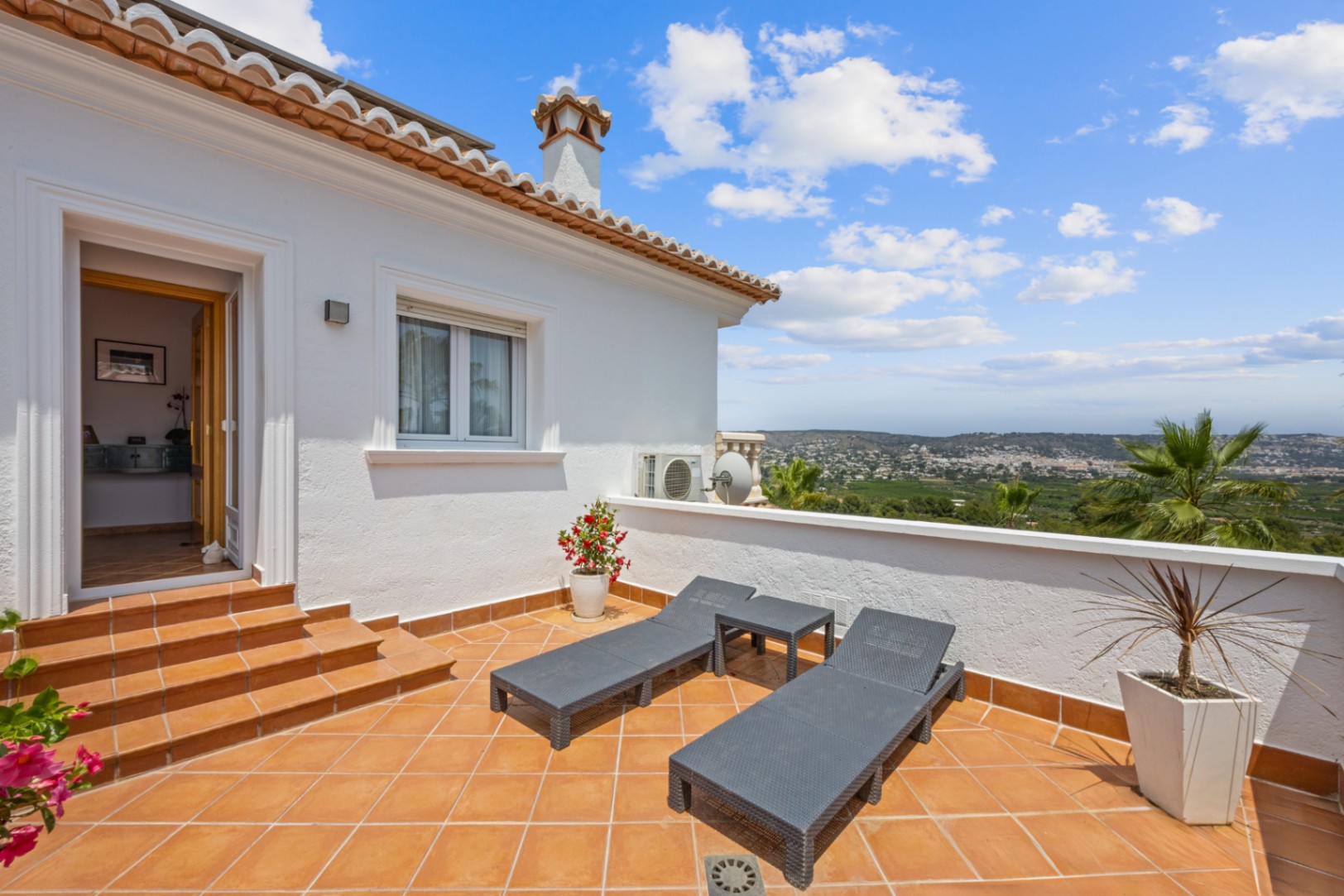 Terraza elevada con suelo de terracota, tumbonas grises y vistas panorámicas al valle. El acceso se realiza mediante escalones desde el interior de la vivienda.