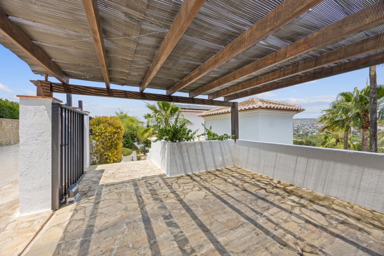 Zona de aparcamiento cubierta con pérgola de madera y cañizo, suelo de piedra natural irregular y vistas despejadas al entorno mediterráneo.