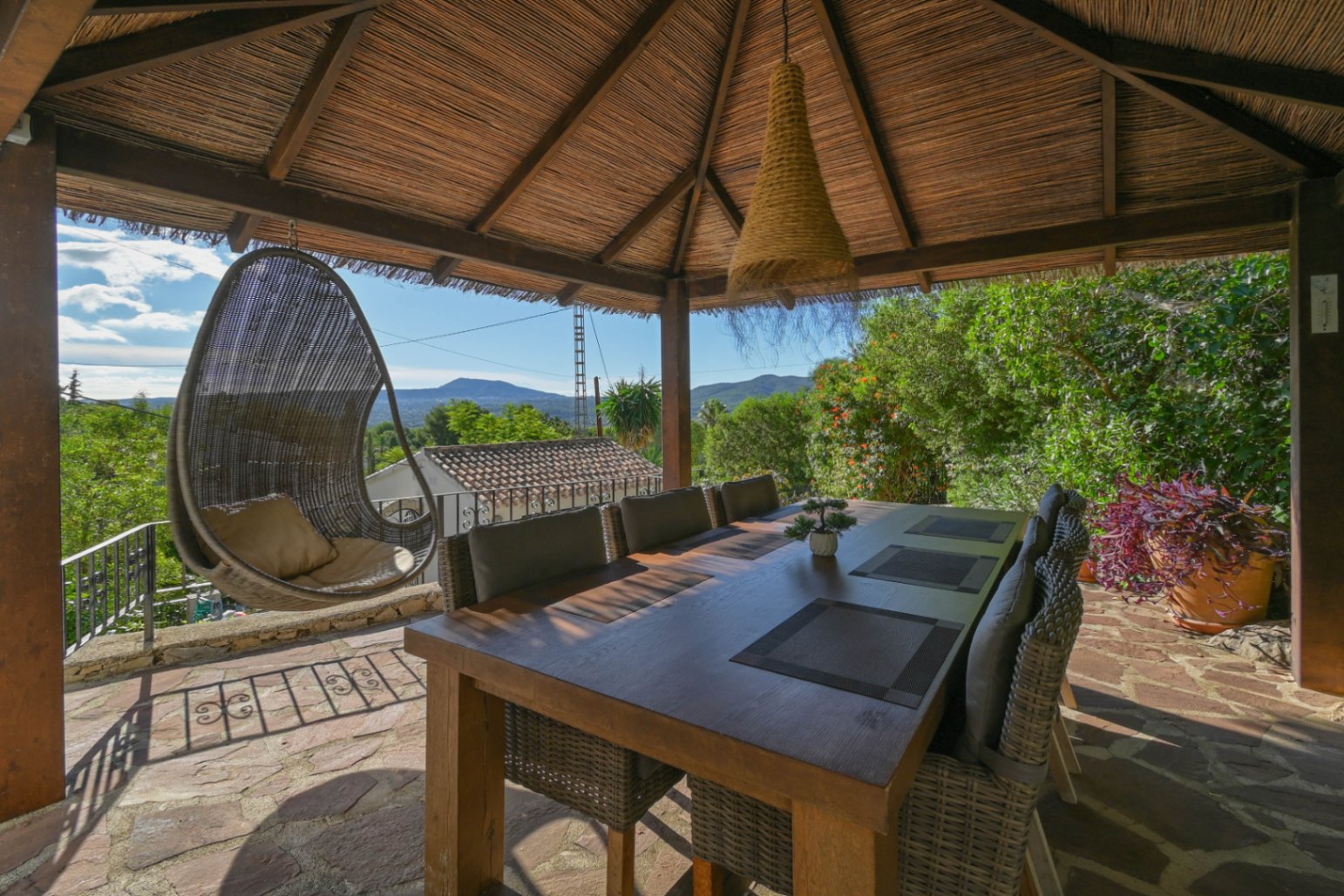 Comedor exterior bajo un cenador de brezo con mesa de madera, sillas de mimbre y un sillón colgante, ofreciendo vistas despejadas a las montañas.