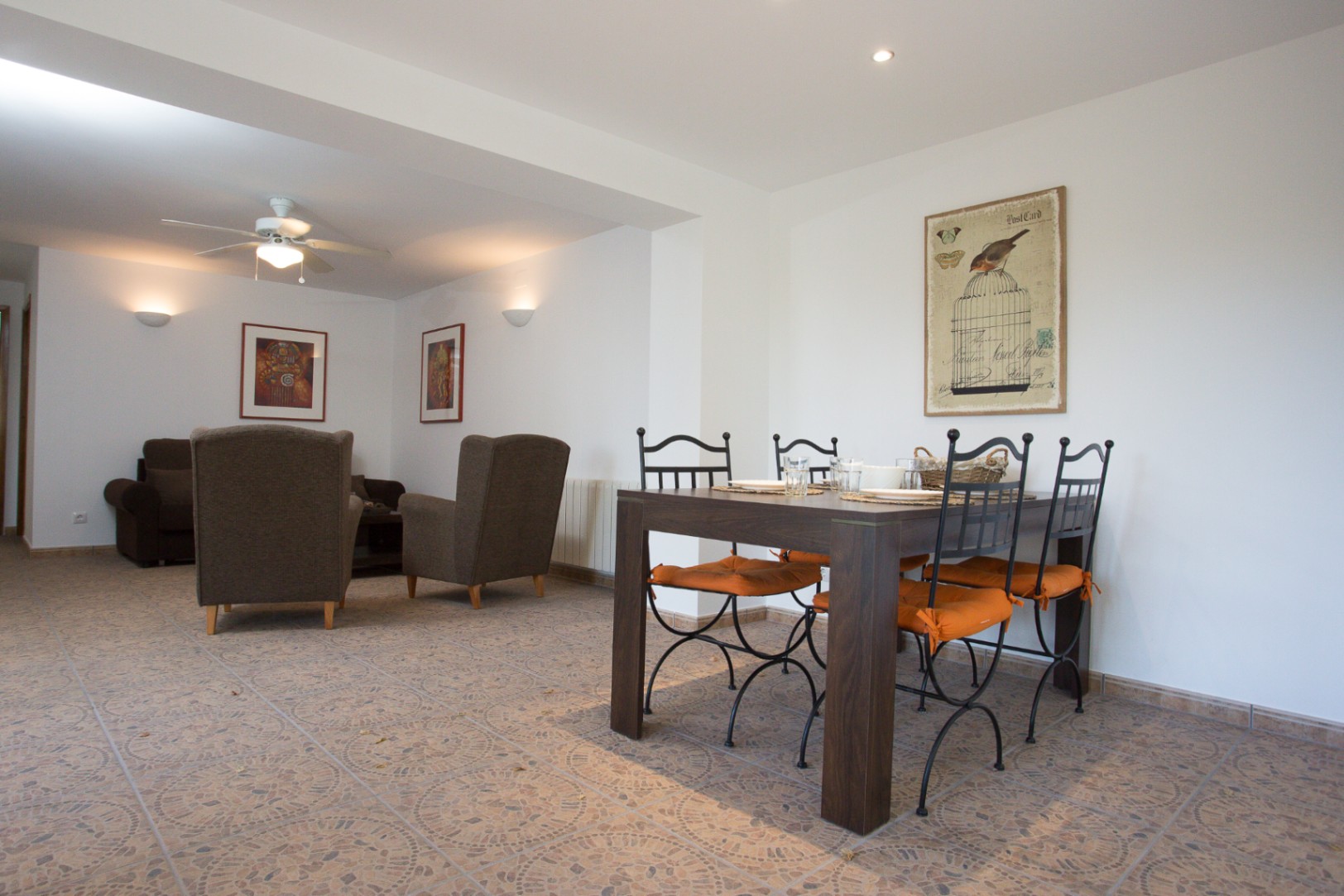 Salón-comedor diáfano con mesa de madera oscura, sillas de forja y cojines naranjas sobre suelo cerámico. Al fondo, zona de estar con butacas y ventilador de techo.