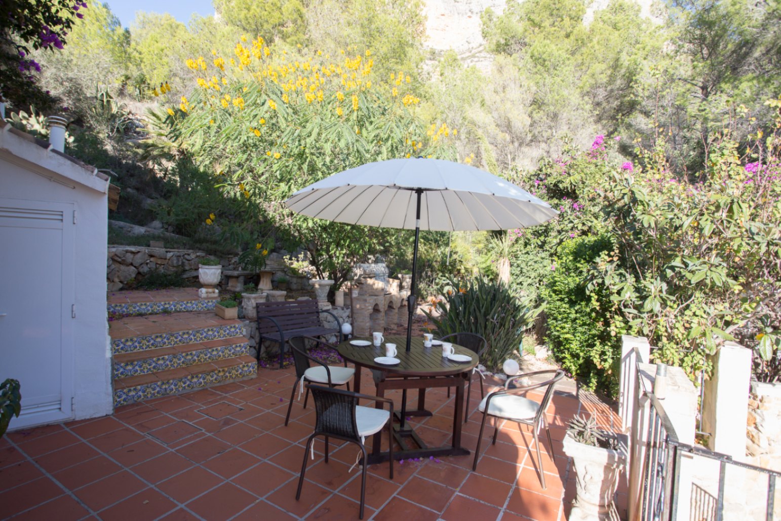 Terraza con suelo de terracota, conjunto de comedor de madera bajo sombrilla blanca y escalones decorados con azulejos tradicionales frente a una ladera boscosa.