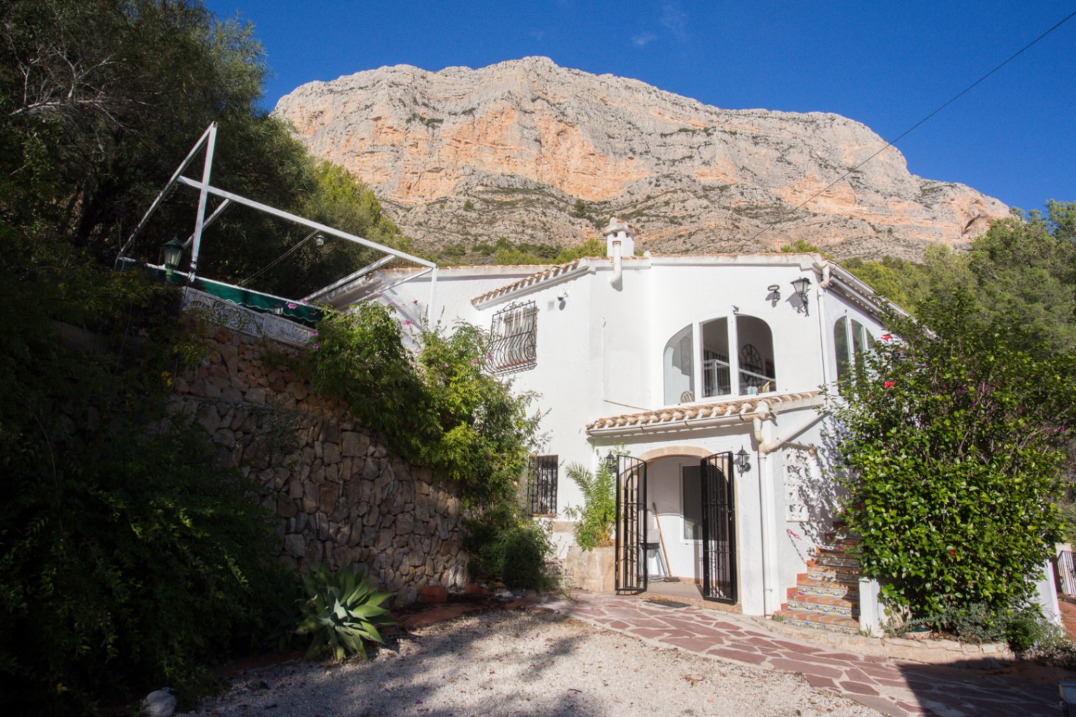 Villa mediterránea blanca con ventanas de arco y muros de piedra, ubicada al pie de una imponente montaña rocosa bajo un cielo despejado.