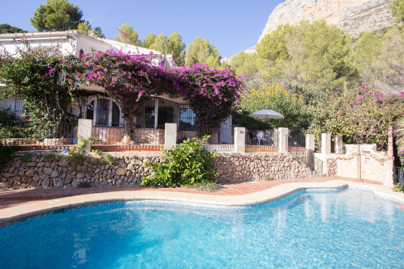 Vista exterior con piscina de forma curva, terraza de terracota y villa blanca adornada con buganvillas frente a un entorno de montaña y piedra natural.