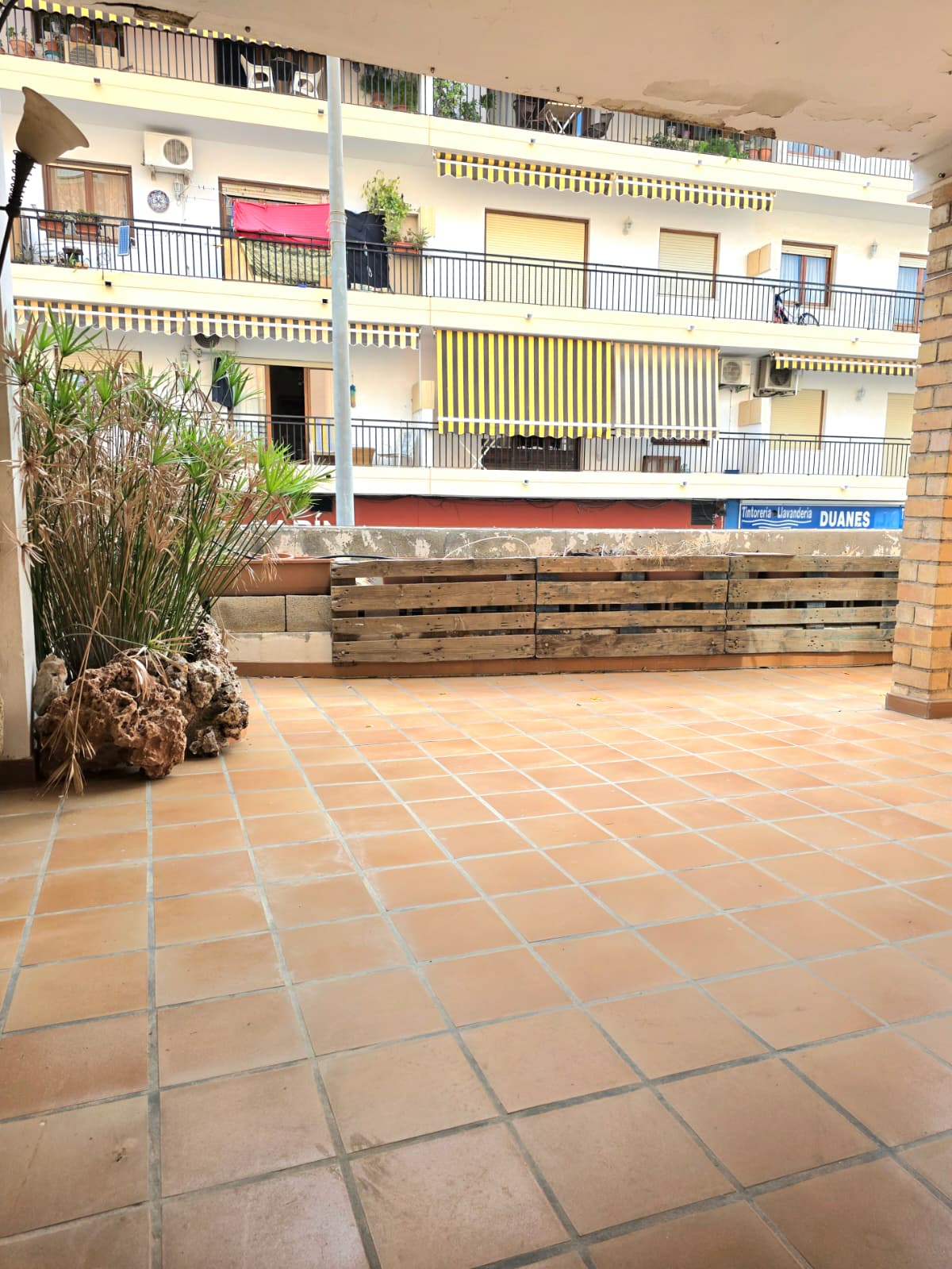 Amplia terraza exterior con suelos de baldosa de terracota, cerramiento de palés de madera y vistas a edificios urbanos con toldos a rayas.