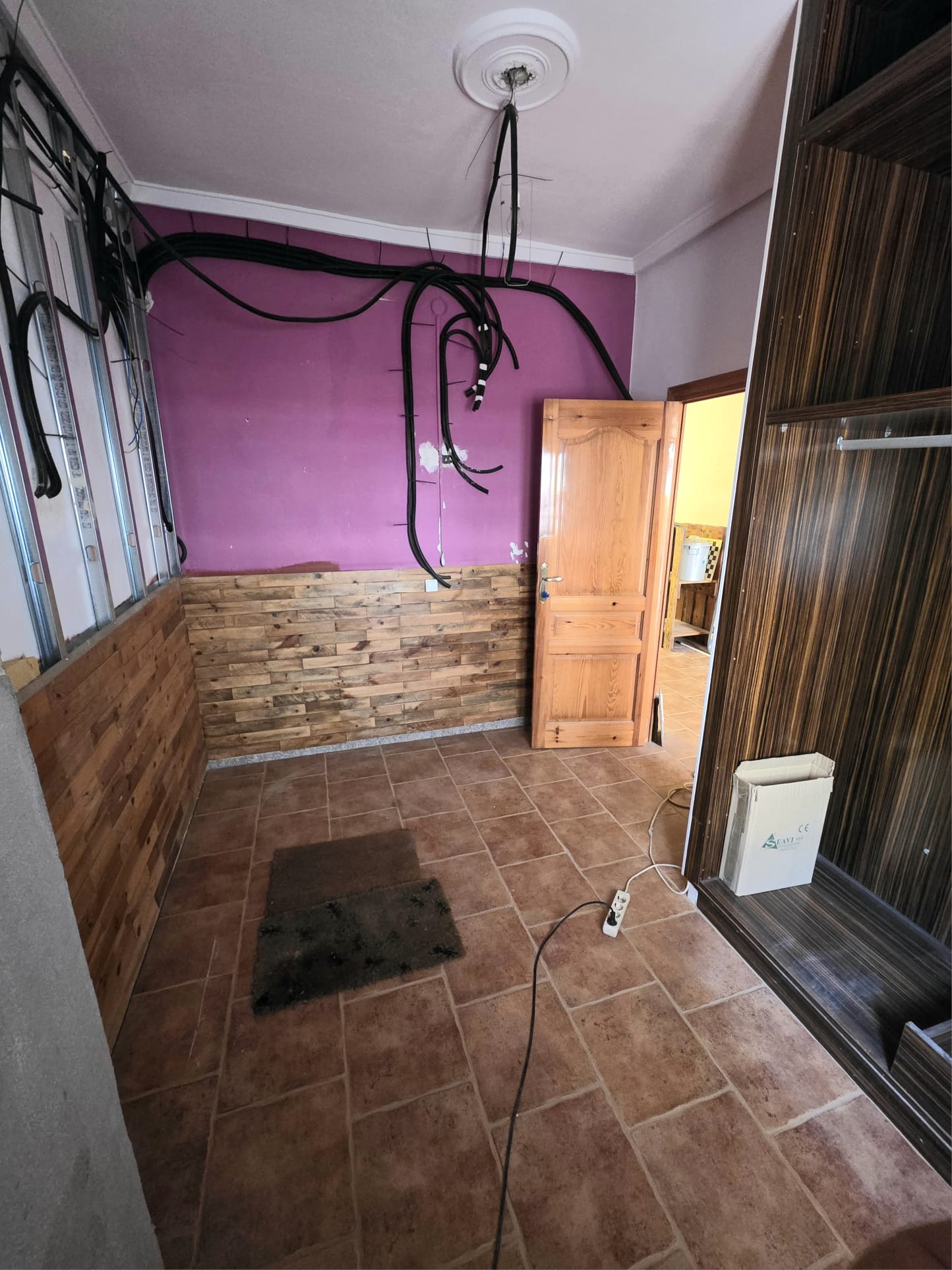 Habitación en reforma con suelo de gres rústico, friso de madera, paredes violetas y cables eléctricos vistos junto a perfiles metálicos.