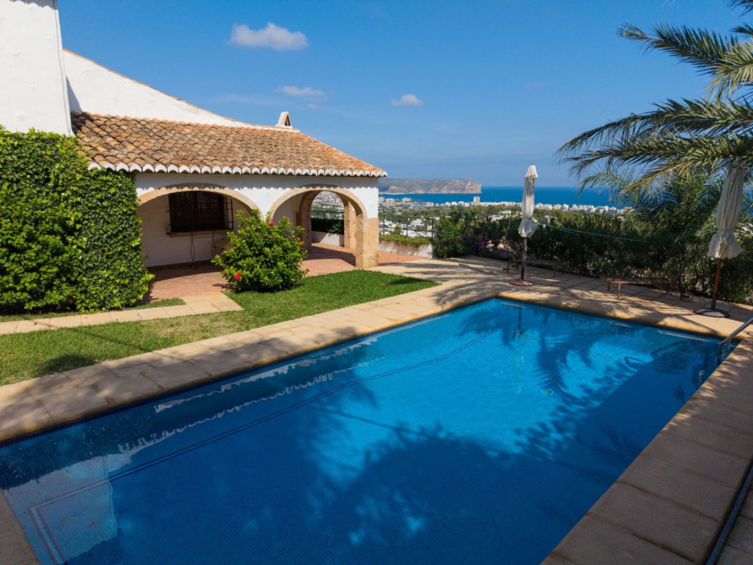 Piscina exterior con borde de piedra junto a una villa mediterránea con porche de arcos y tejado de terracota, con vistas panorámicas al mar y la costa.