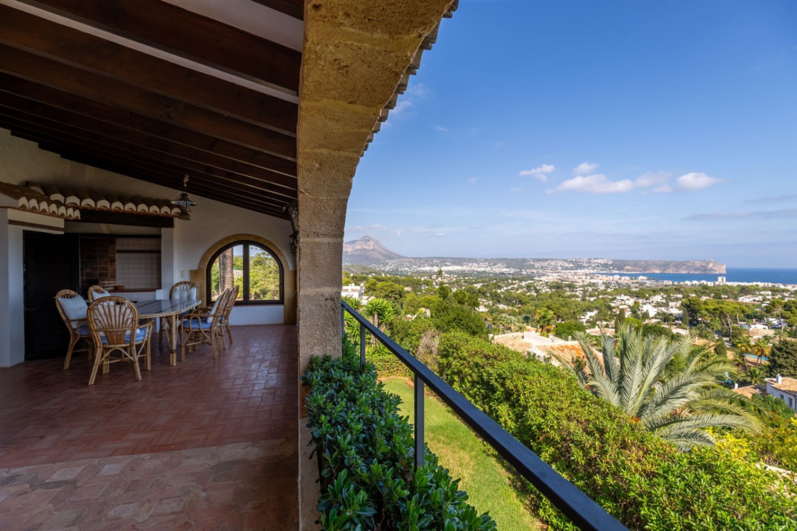 Terraza cubierta con vigas de madera y suelo de terracota, zona de comedor exterior y vistas panorámicas al valle, el mar y la montaña.