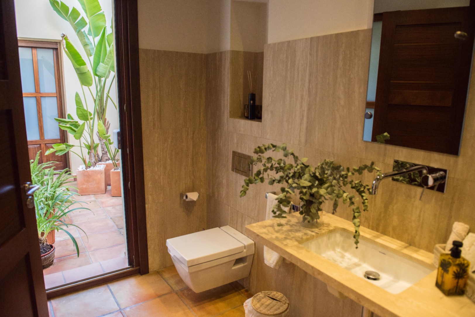 Baño moderno con revestimiento de travertino, inodoro suspendido y acceso directo a un patio privado con suelos de terracota y plantas tropicales.