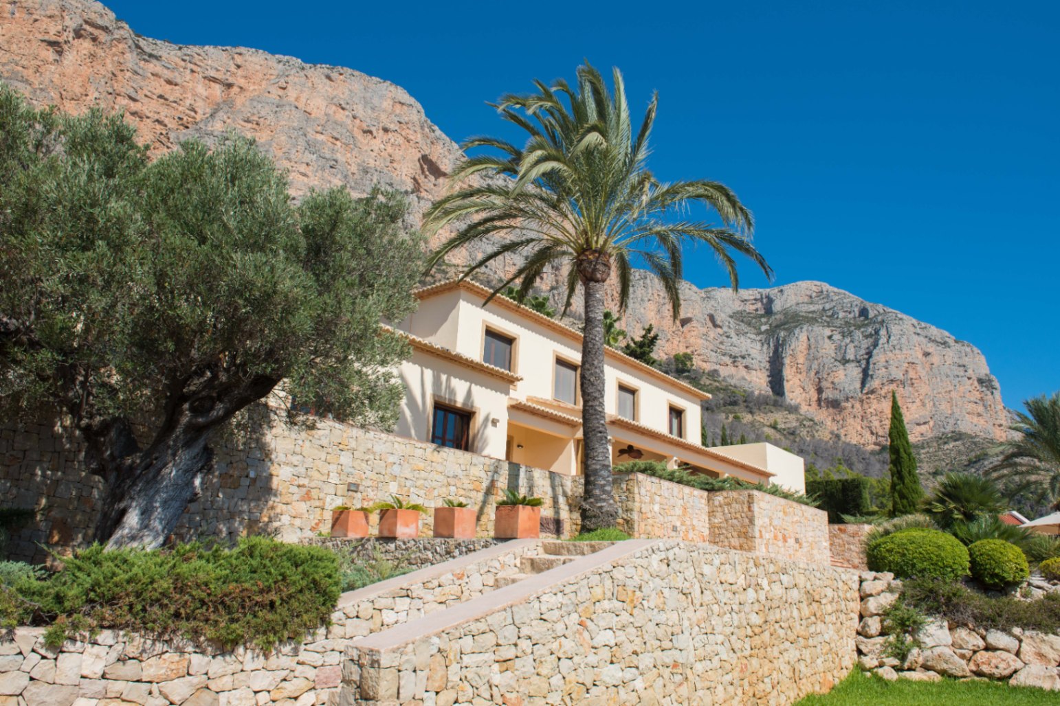 Fachada de una villa mediterránea con muros de piedra natural, un olivo centenario y una palmera, situada bajo un imponente macizo rocoso y cielo despejado.