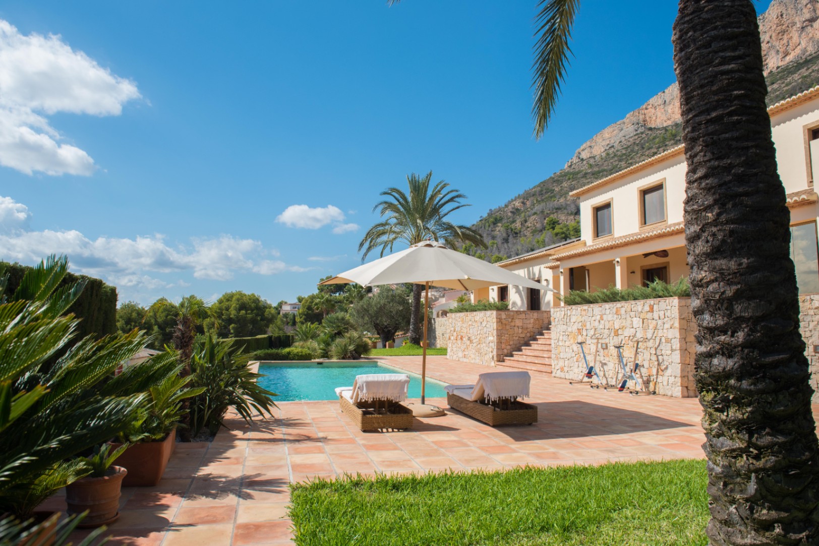 Terraza de baldosas cerámicas con piscina rectangular, tumbonas de mimbre y muros de piedra seca. El espacio cuenta con vegetación mediterránea y vistas a la ladera de la montaña.