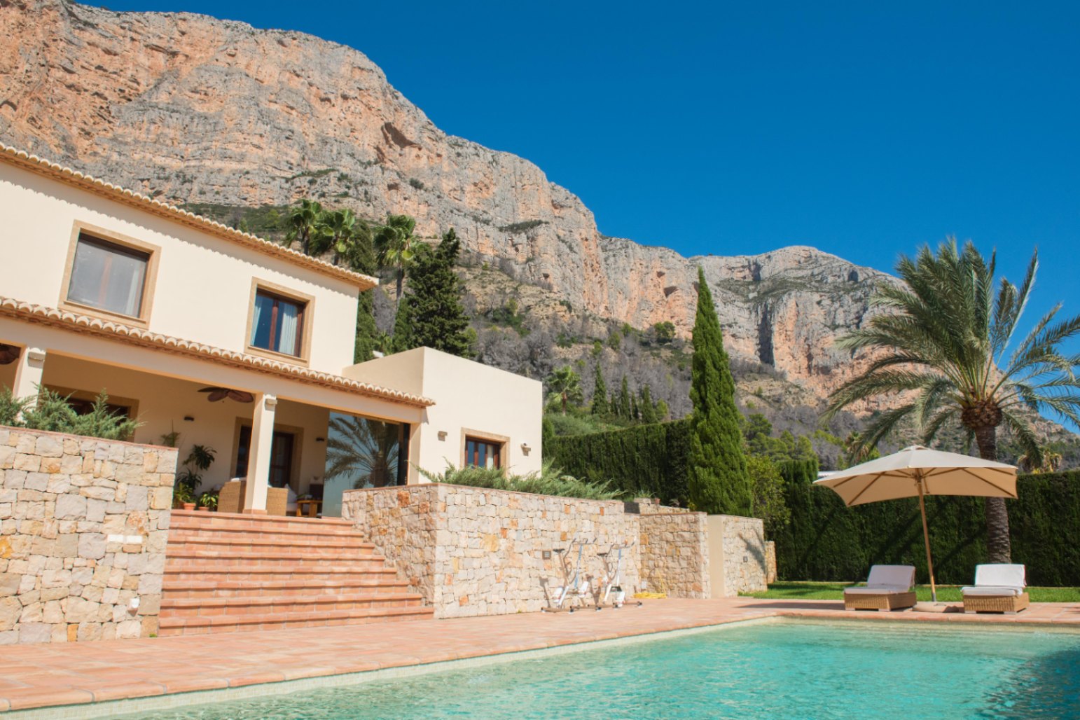 Vista exterior con piscina turquesa, terraza de terracota y muros de piedra natural bajo una imponente montaña de relieve escarpado y vegetación mediterránea.