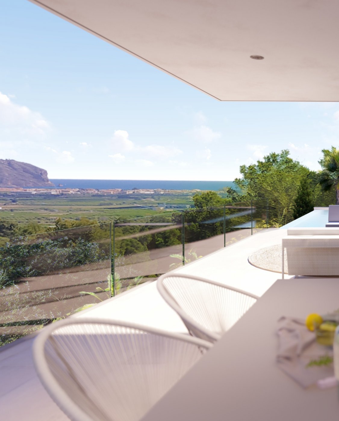 Terraza elevada con barandilla de cristal y vistas panorámicas a la costa mediterránea y las montañas. Incluye mobiliario de diseño en tonos blancos y suelos de gres claro.