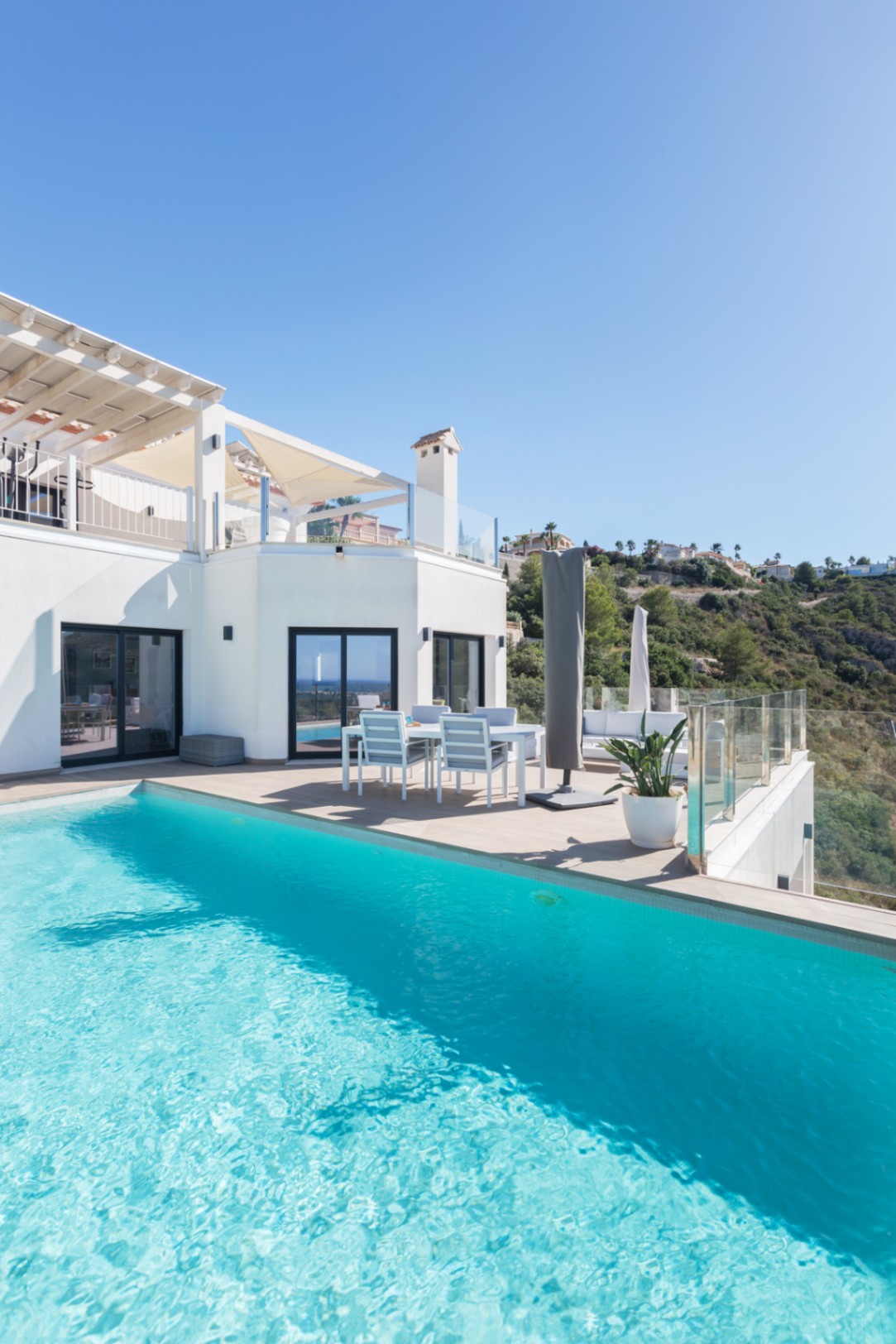 Piscina infinita de aguas turquesas junto a una terraza de gres cerámico con comedor exterior y barandillas de cristal con vistas al valle.