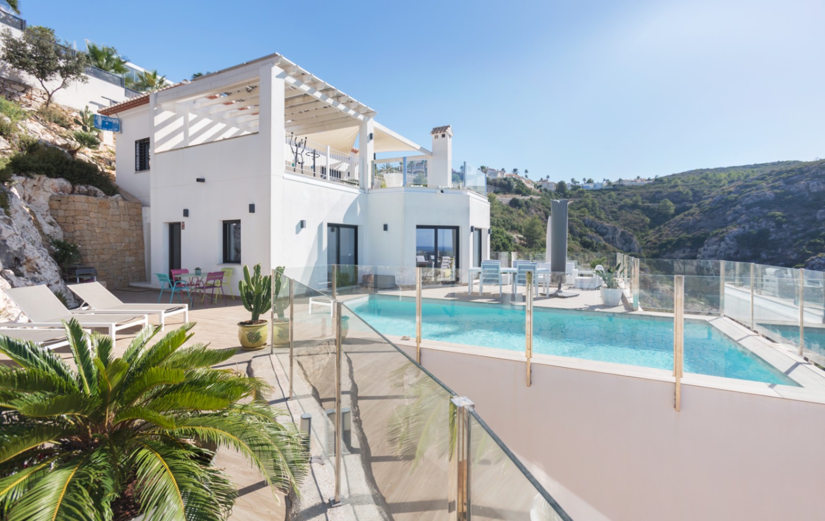 Vista exterior de una villa blanca de varios niveles con piscina, barandillas de cristal, terrazas solárium y vistas panorámicas a la montaña.
