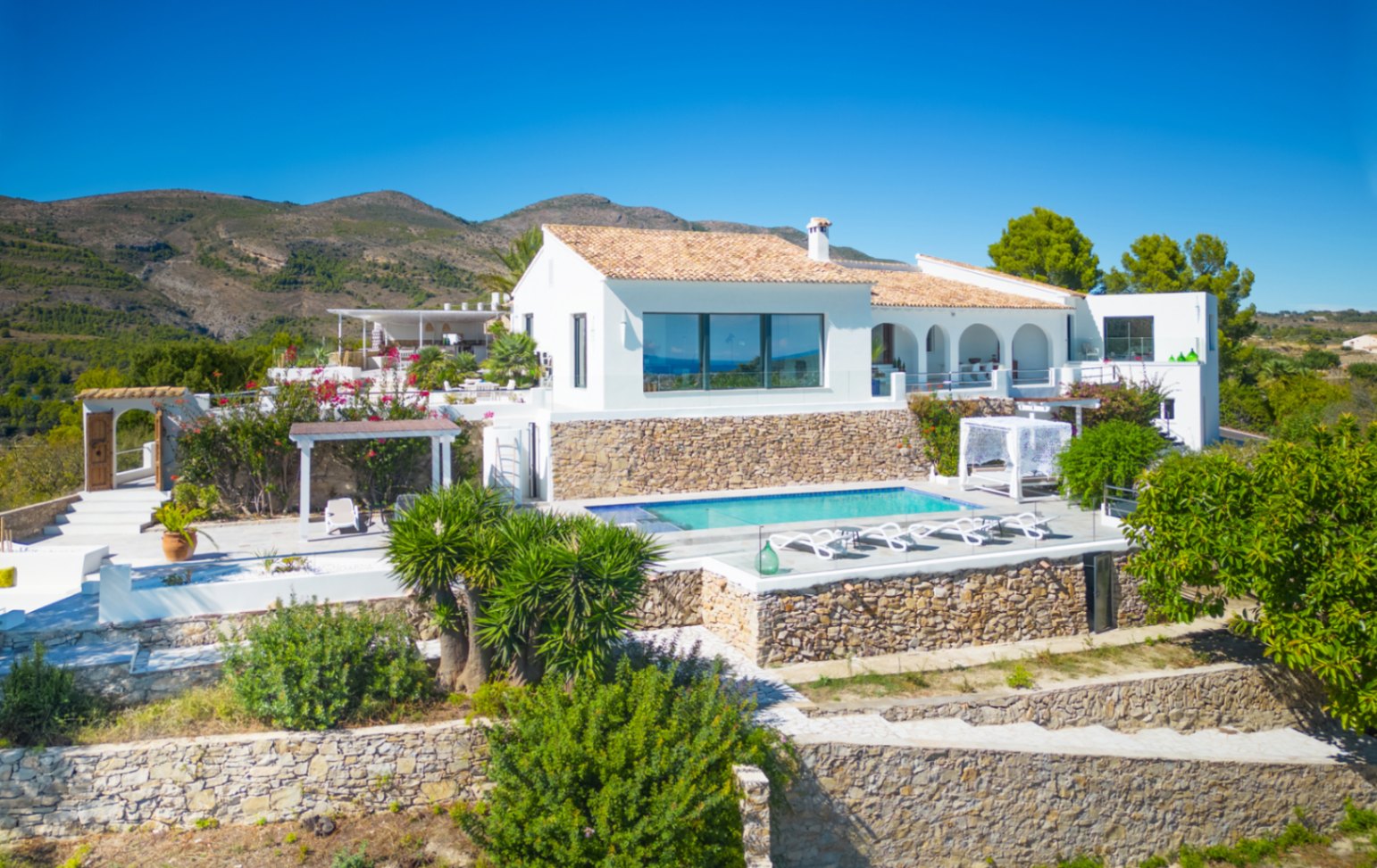 Fachada de villa mediterránea blanca con piscina, muros de mampostería y porches arqueados, integrada en un entorno montañoso.