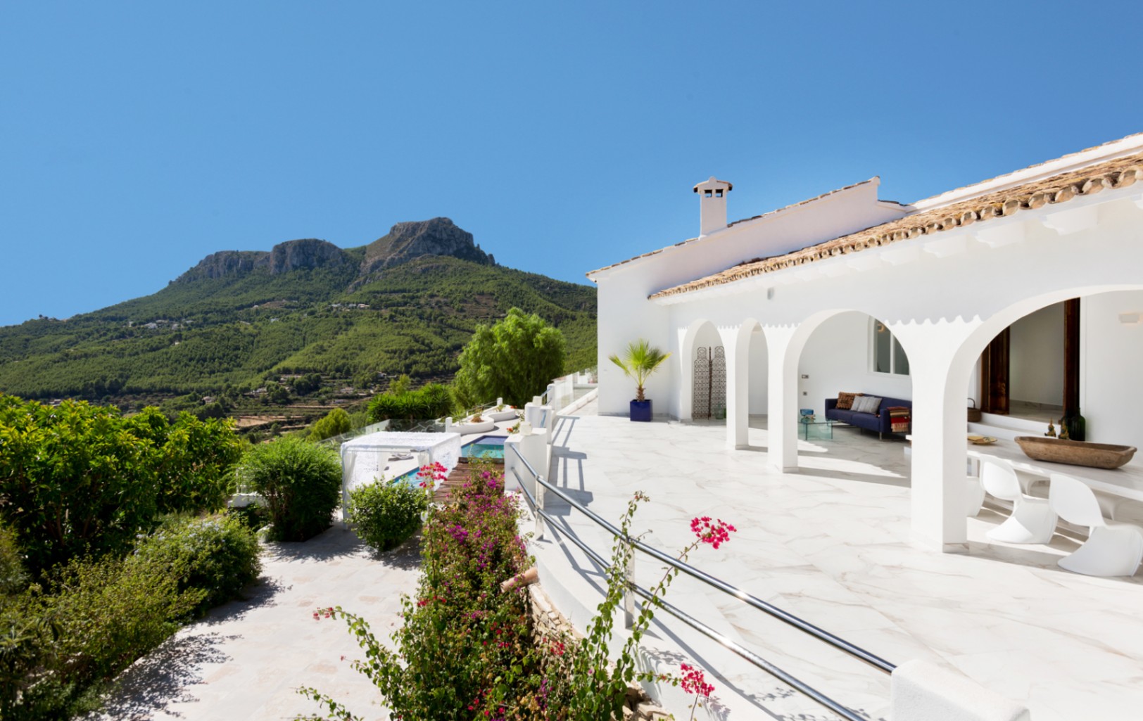 Villa mediterránea blanca con terraza de arcos y suelo de mármol, zona de estar exterior y amplias vistas al exuberante paisaje montañoso.