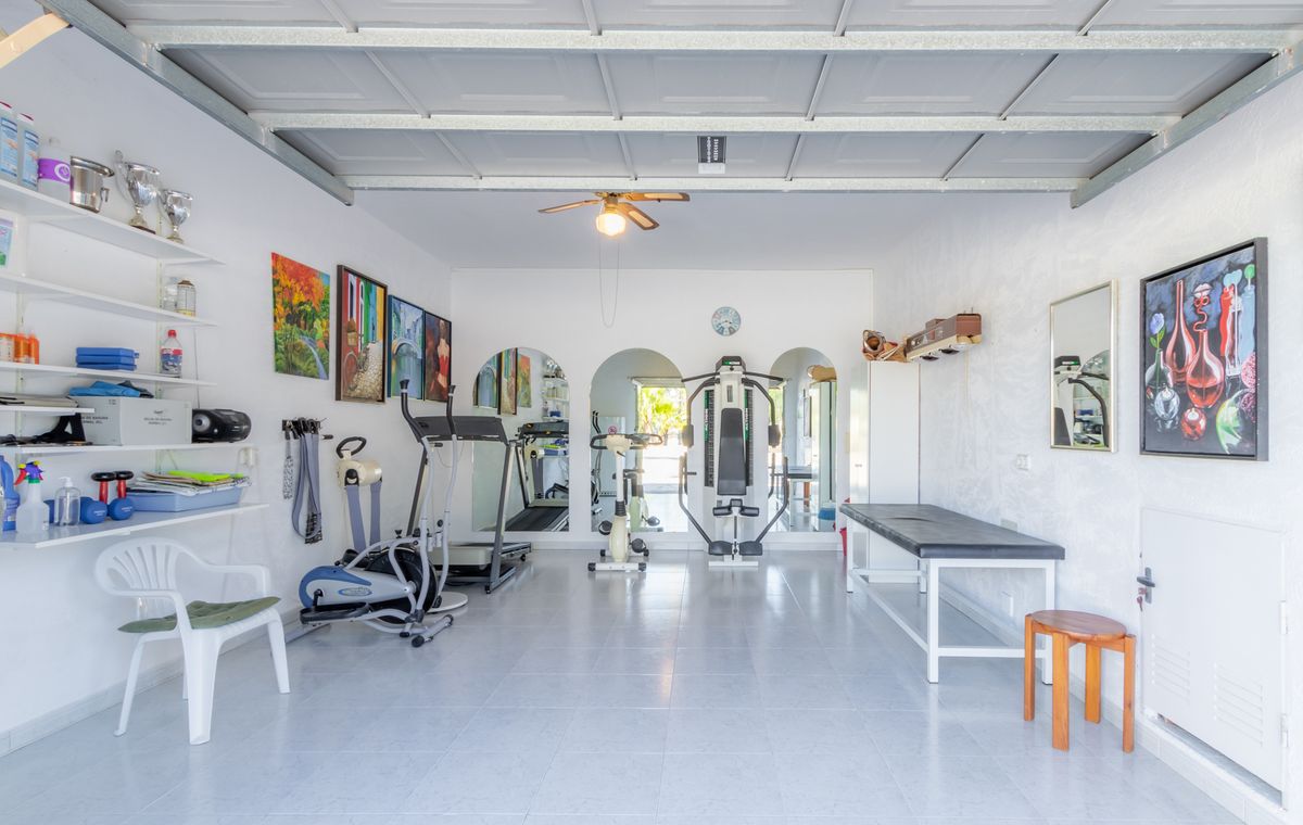 Gimnasio privado en garaje reformado con suelos cerámicos, equipado con máquinas de cardio, estación de pesas, camilla de tratamiento y espejos arqueados.