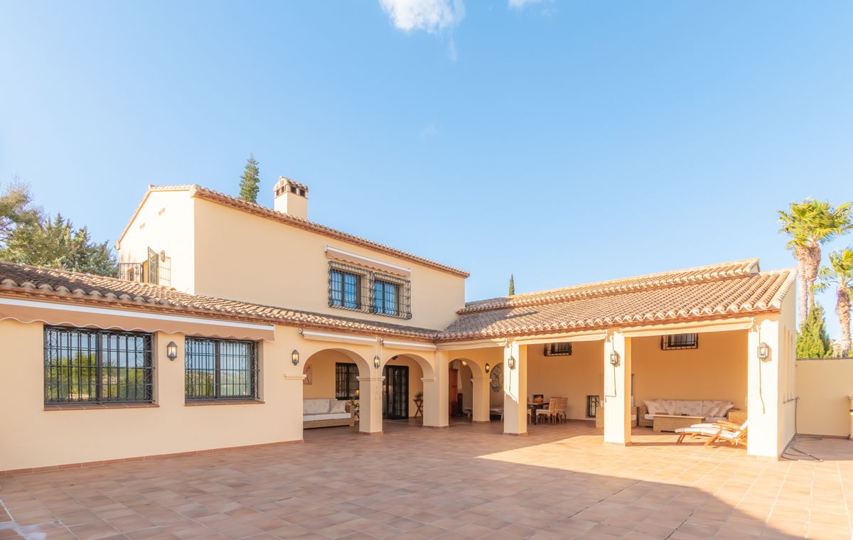 Vista exterior de una villa mediterránea con una amplia terraza de baldosas de terracota, porches con arcos, zonas de estar al aire libre y fachada de color crema.