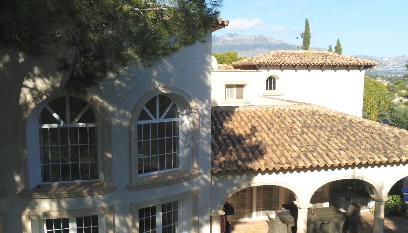 Fachada de villa mediterránea con tejados de teja cerámica, ventanales de arco y porche cubierto con vistas a las montañas.
