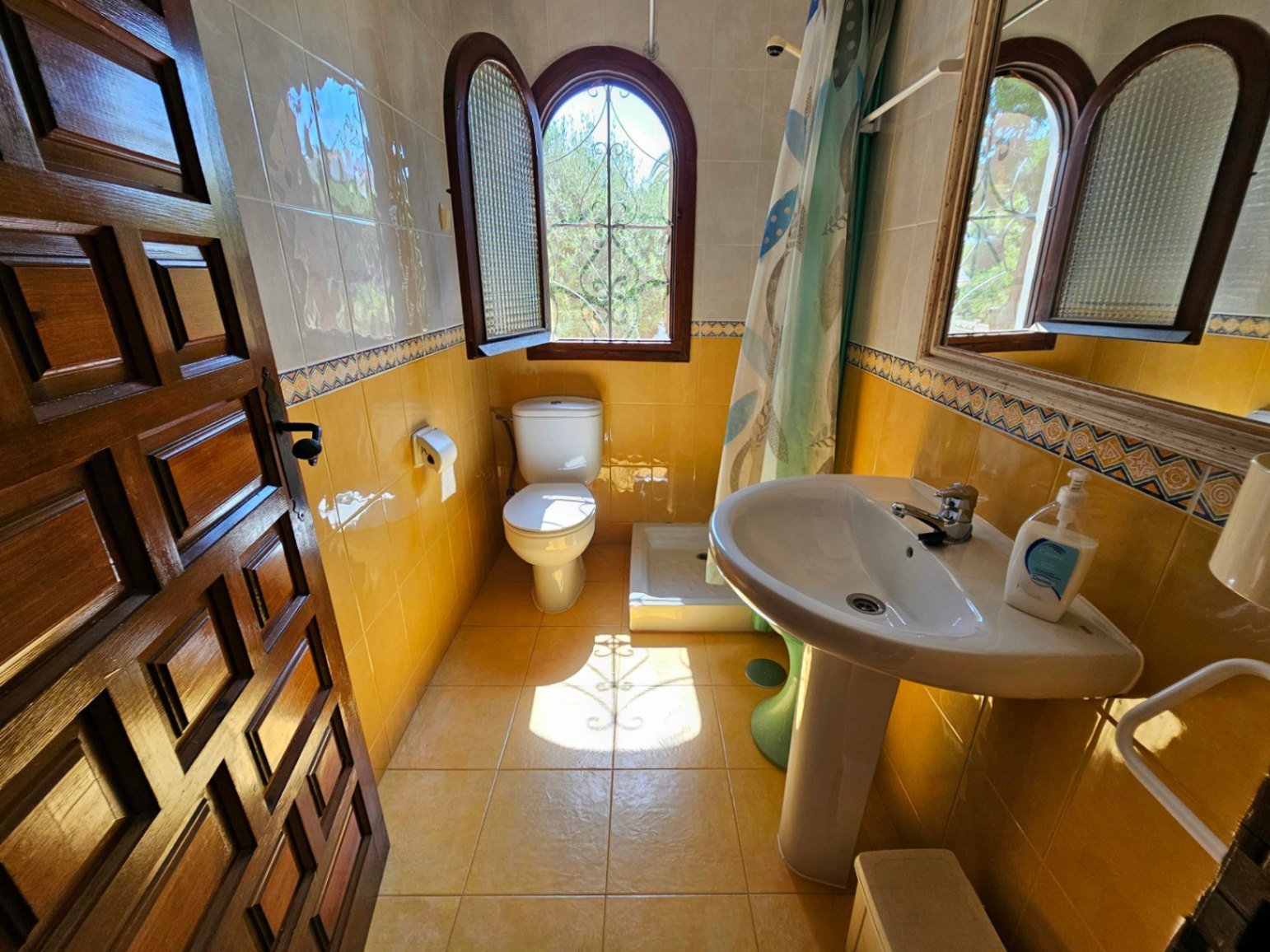Baño de estilo tradicional con azulejos amarillos, lavabo de pedestal blanco y ventana de arco en madera con rejería decorativa.