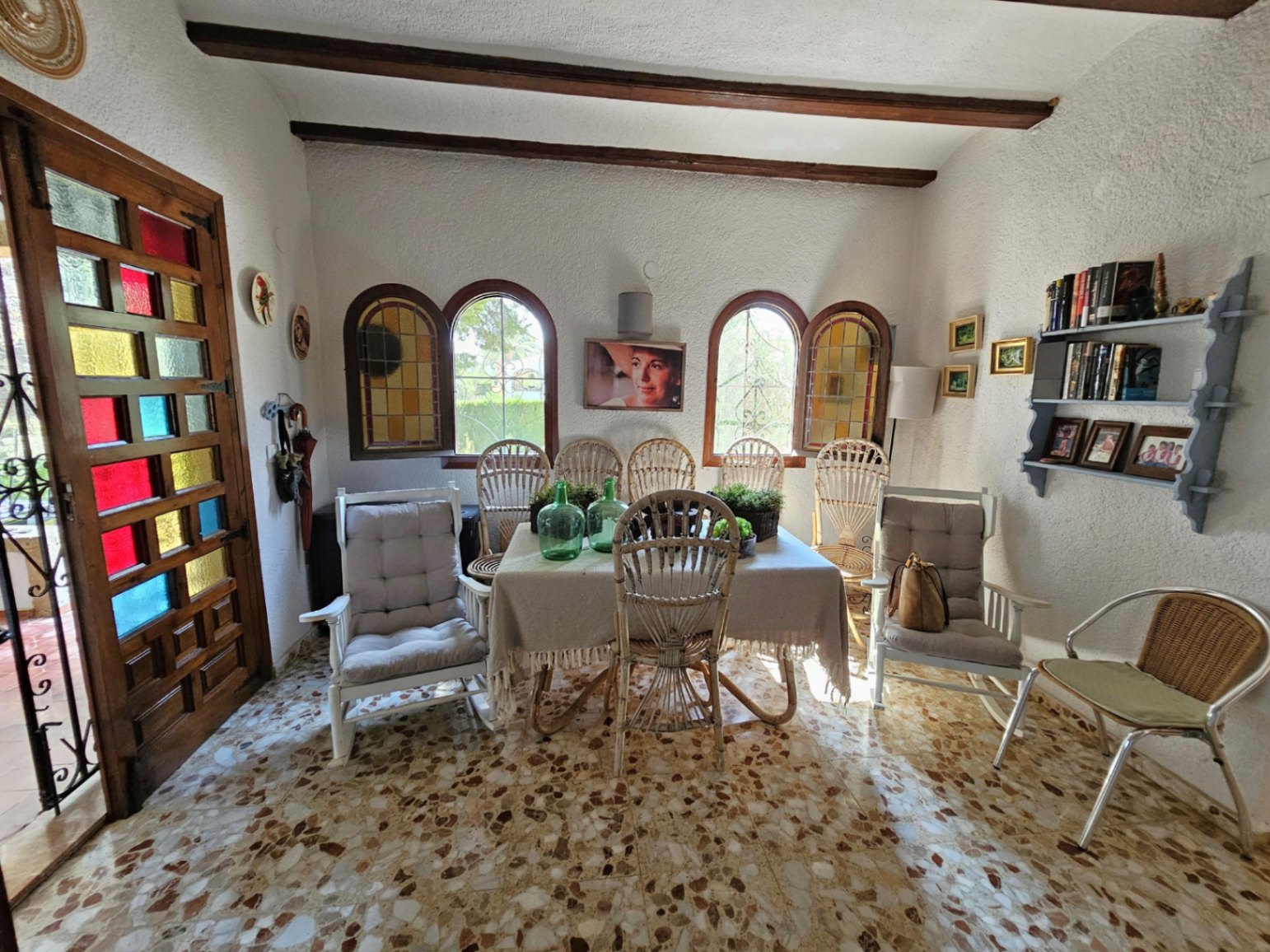 Comedor rústico con suelo de terrazo, vigas de madera vista y ventanas de arco. Destaca la puerta de madera con paneles de cristal tintado de colores.