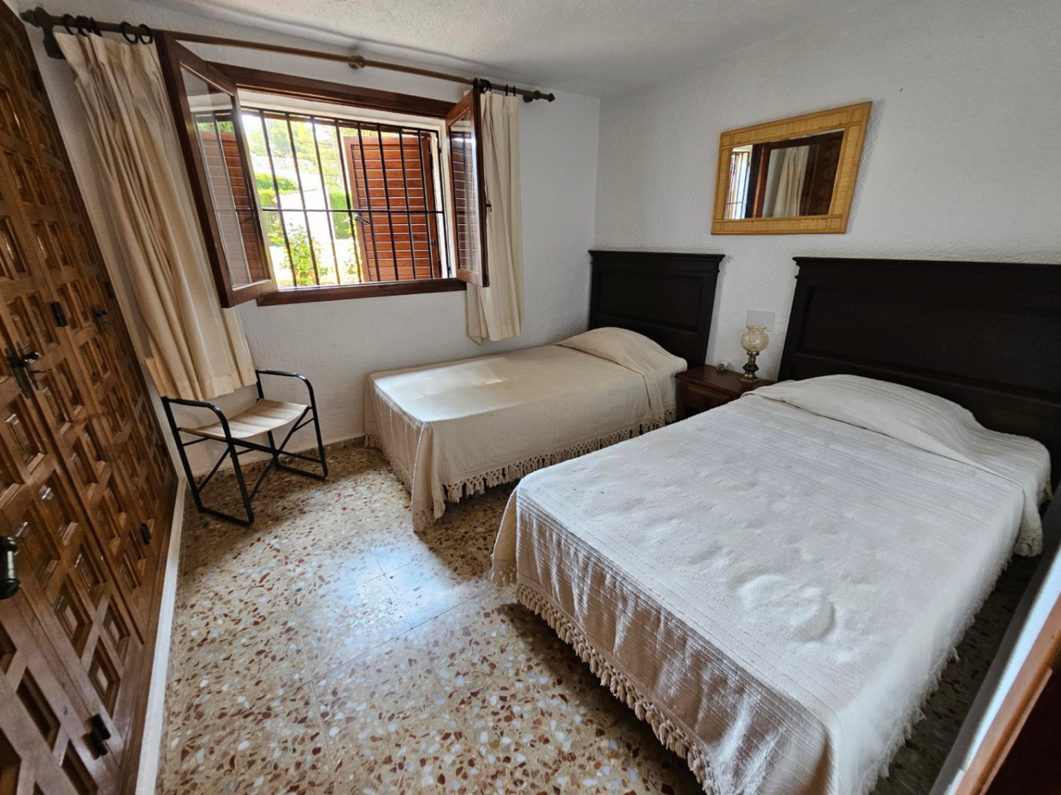 Dormitorio con dos camas individuales, cabeceros de madera oscura, suelo de terrazo original y ventana de madera con contraventanas y rejas de seguridad.