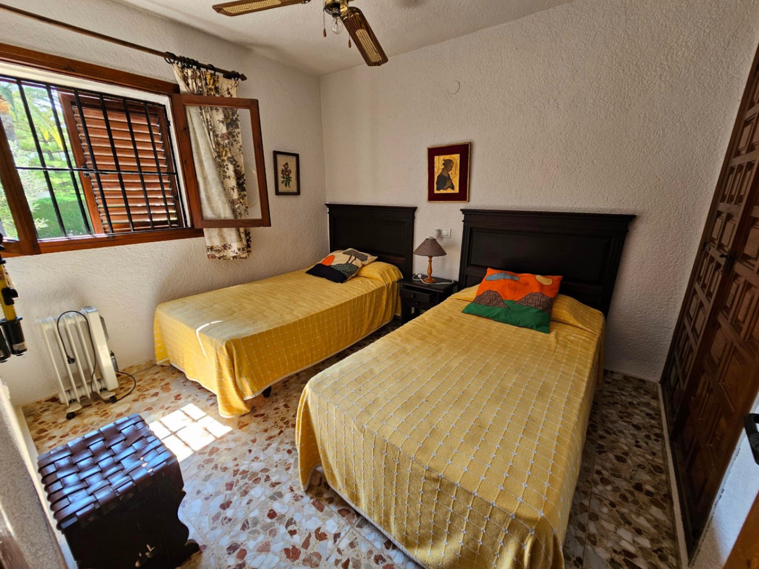 Dormitorio con dos camas individuales, cabeceros de madera oscura y suelo de terrazo original. Dispone de ventilador de techo y ventana con rejas de seguridad.