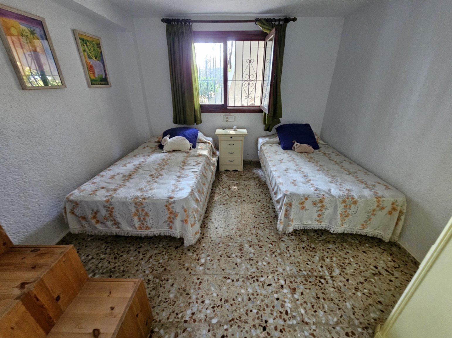 Dormitorio con dos camas individuales, suelos de terrazo y mesita de noche. Incluye ventana con rejas de seguridad, cortinas verdes y paredes de acabado rugoso.