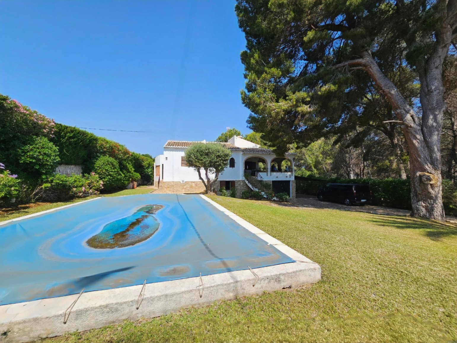 Exterior de una villa blanca con porche arqueado y base de piedra, rodeada de un jardín con césped, pinos maduros y una piscina con cubierta protectora.