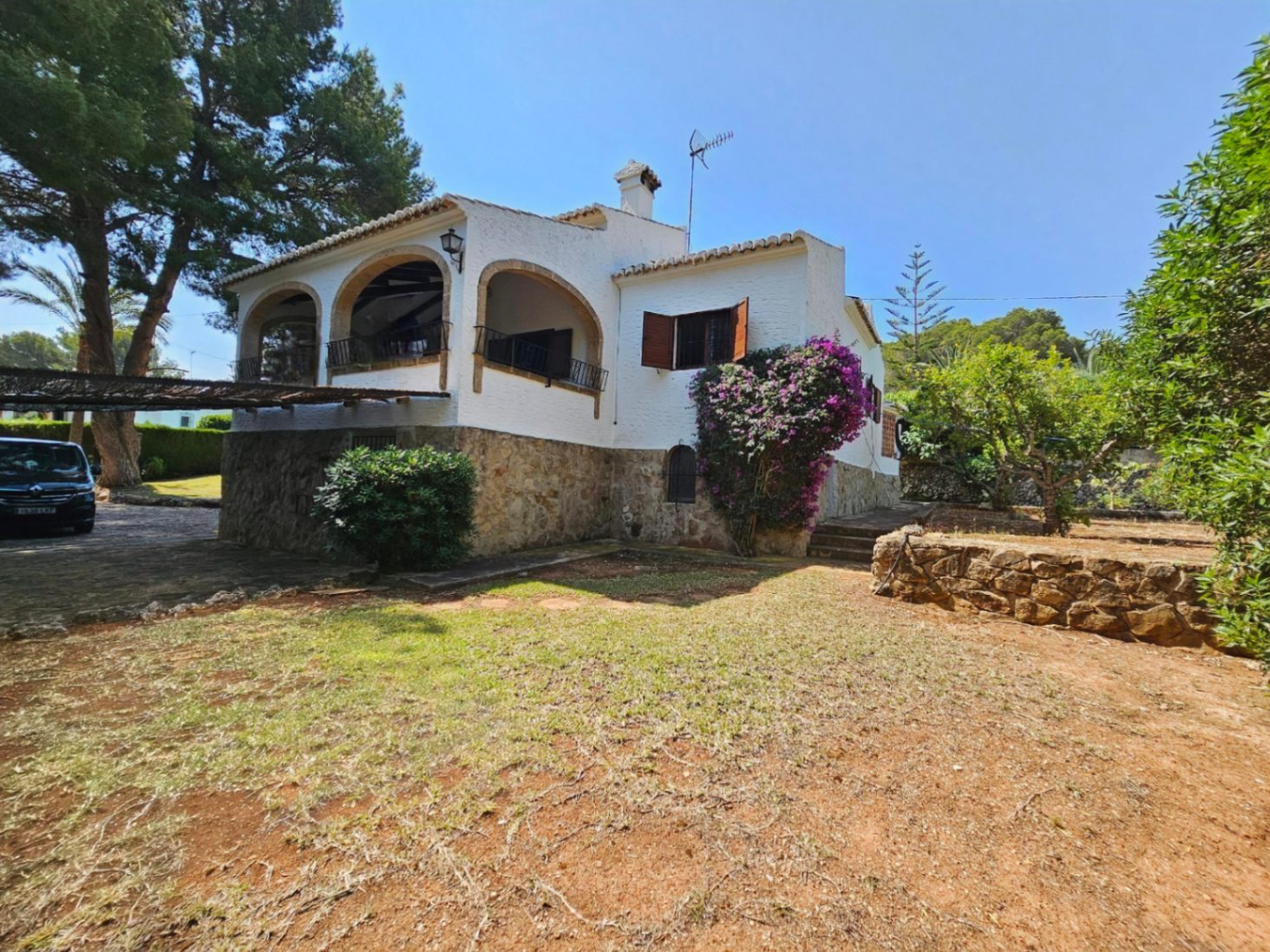 Exterior de villa mediterránea con base de piedra natural, terraza arqueada con barandillas de forja y buganvilla sobre muros blancos.