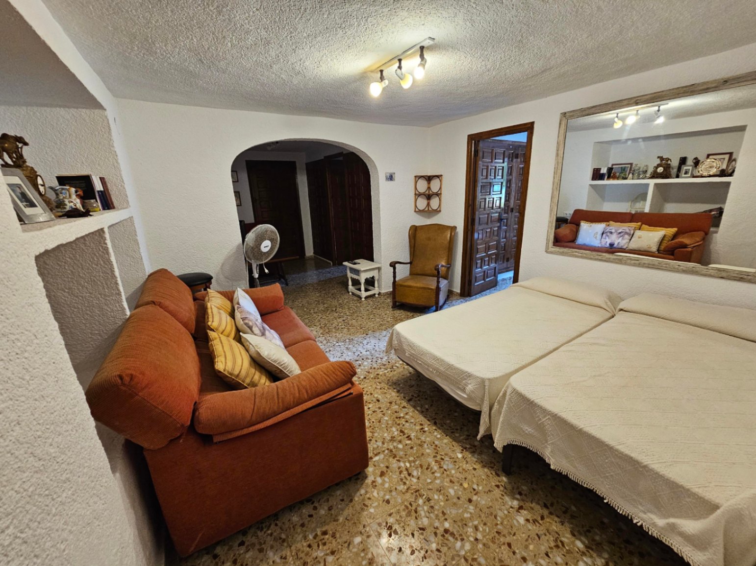 Habitación con suelo de terrazo, dos camas individuales, sofá naranja y hornacinas de obra. Destaca el arco de paso y un gran espejo con marco de madera.