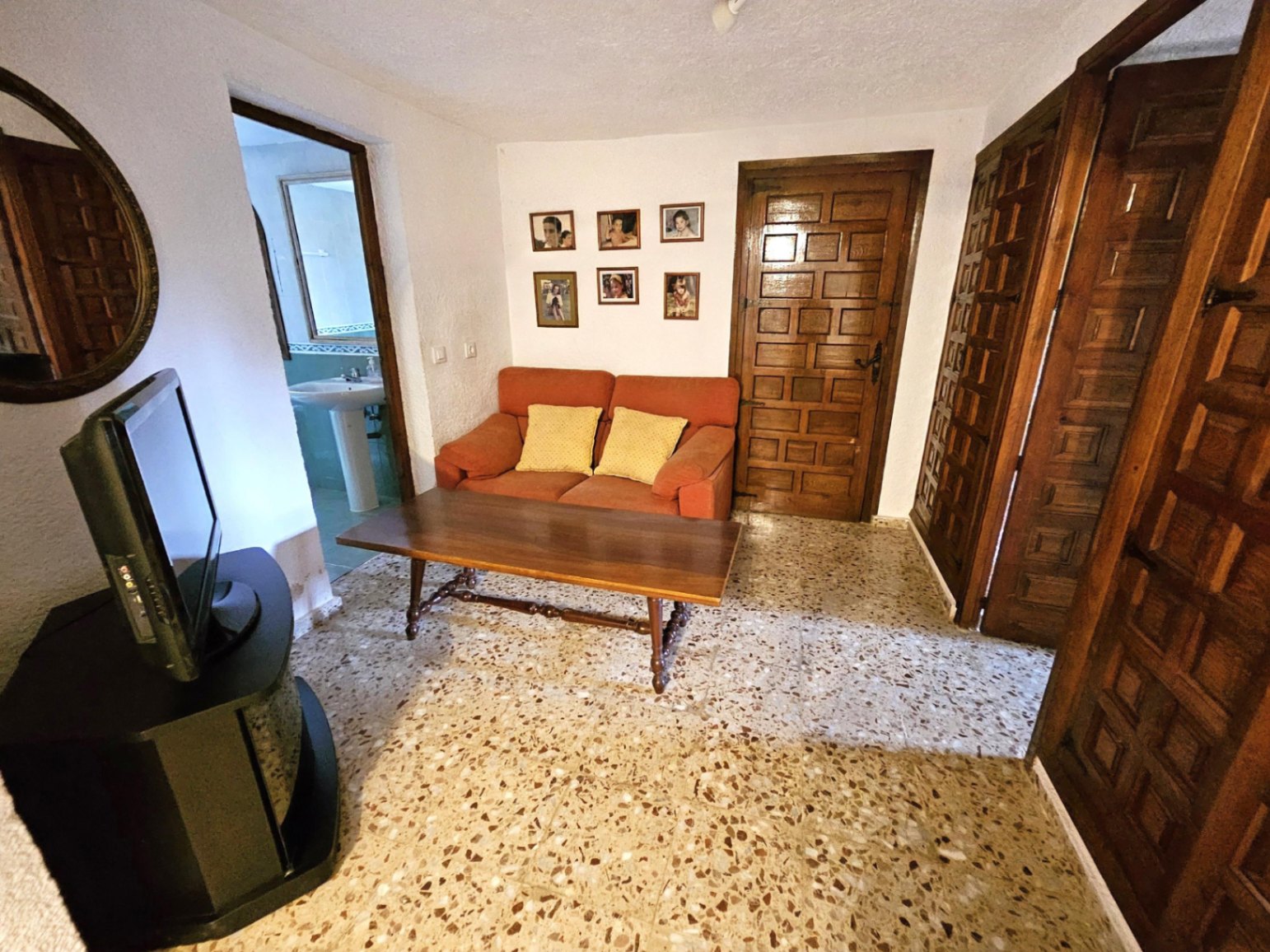 Sala de estar con sofá terracota, puertas de madera tallada y suelo de terrazo. Se observa el acceso a un baño con lavabo de pedestal.