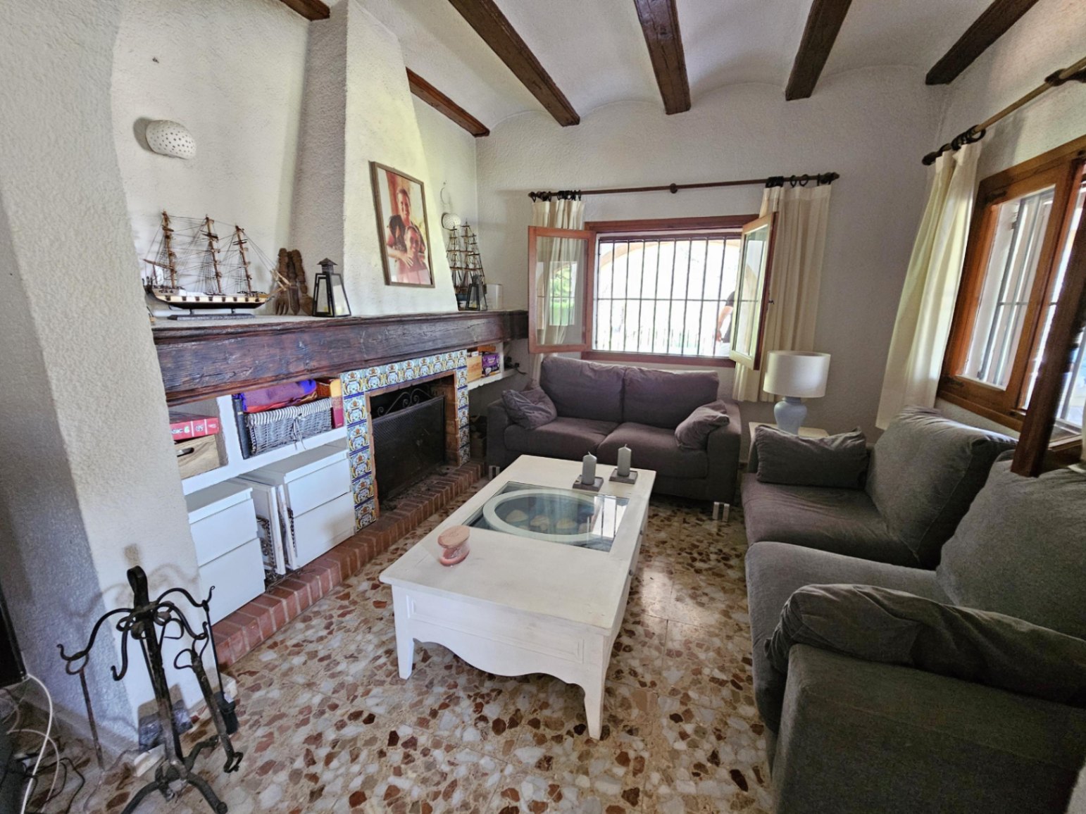Salón con vigas de madera a la vista, chimenea rústica con detalles cerámicos y suelo de terrazo tradicional bajo luz natural.