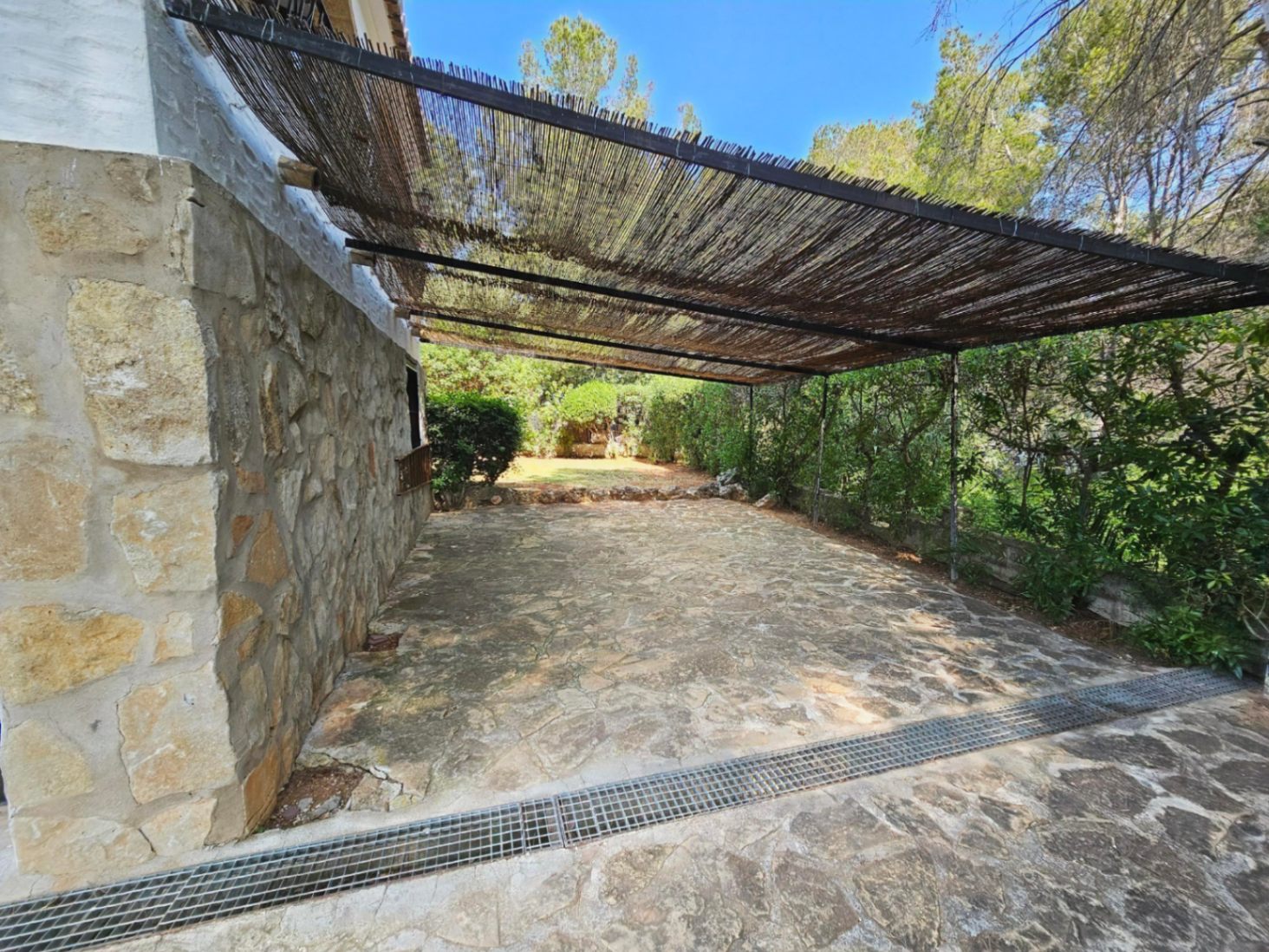 Terraza con pavimento de piedra irregular y muro de mampostería, protegida por una pérgola de cañizo rodeada de vegetación natural.