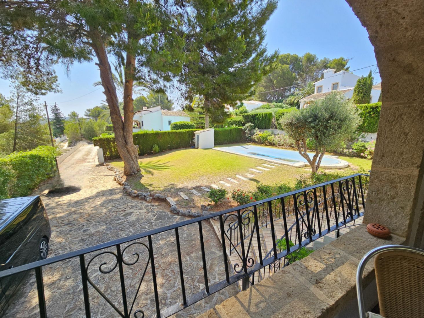 Vista desde una terraza de piedra con barandilla de forja hacia el jardín con césped, piscina y camino empedrado rodeado de pinos mediterráneos.