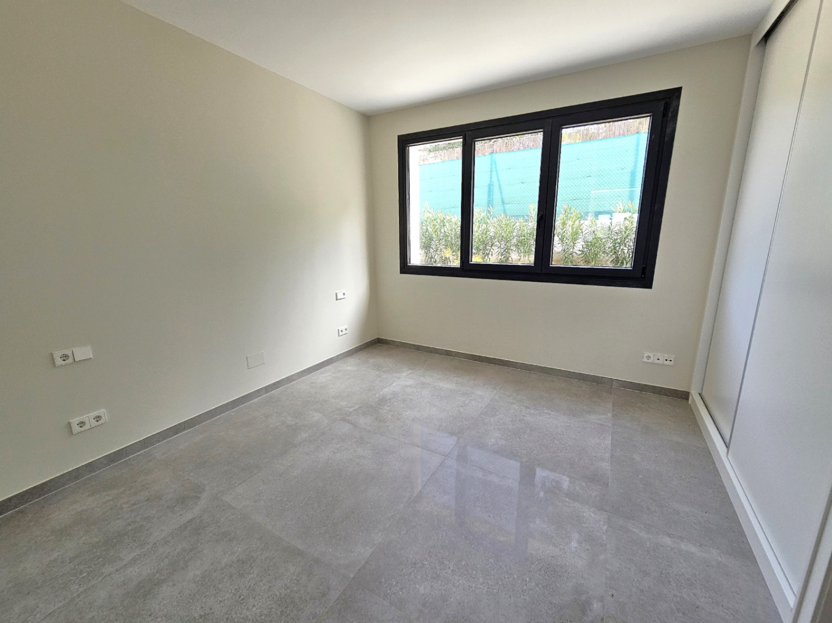 Dormitorio luminoso con suelo de gres porcelánico y ventana de triple panel con marco negro.