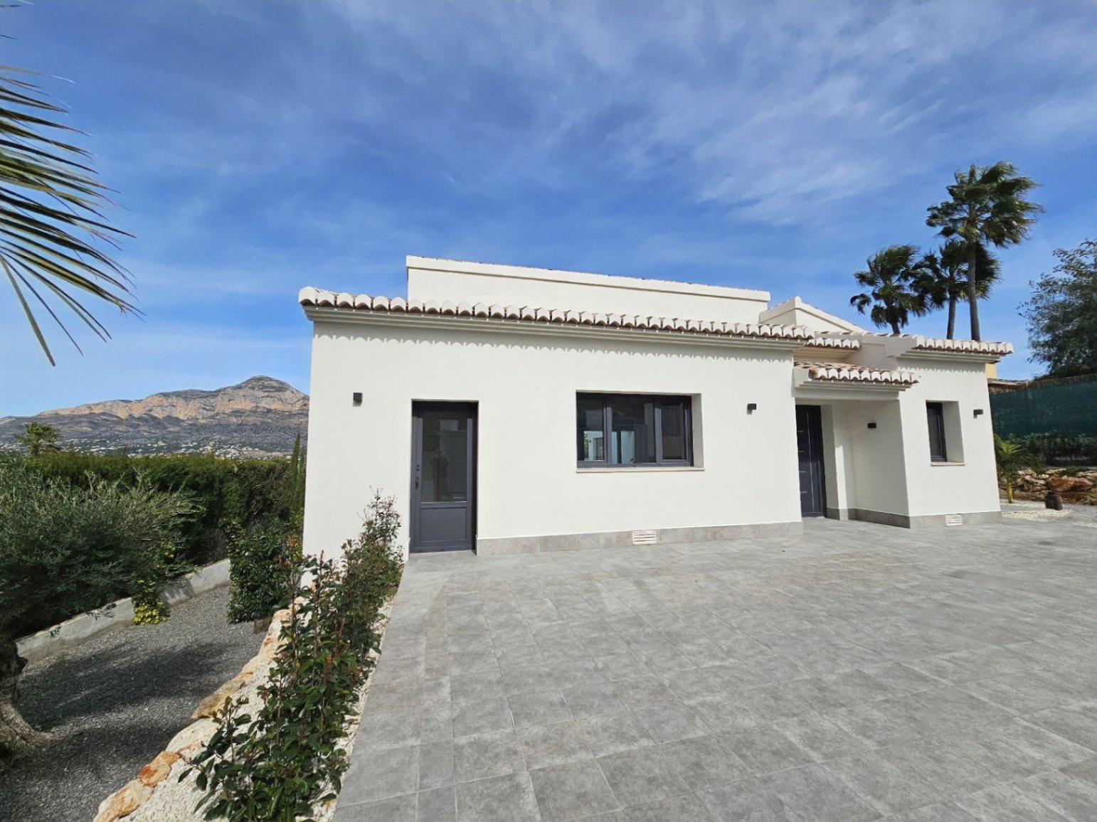Vista exterior de la villa con fachada blanca, tejas y acceso pavimentado en gris.