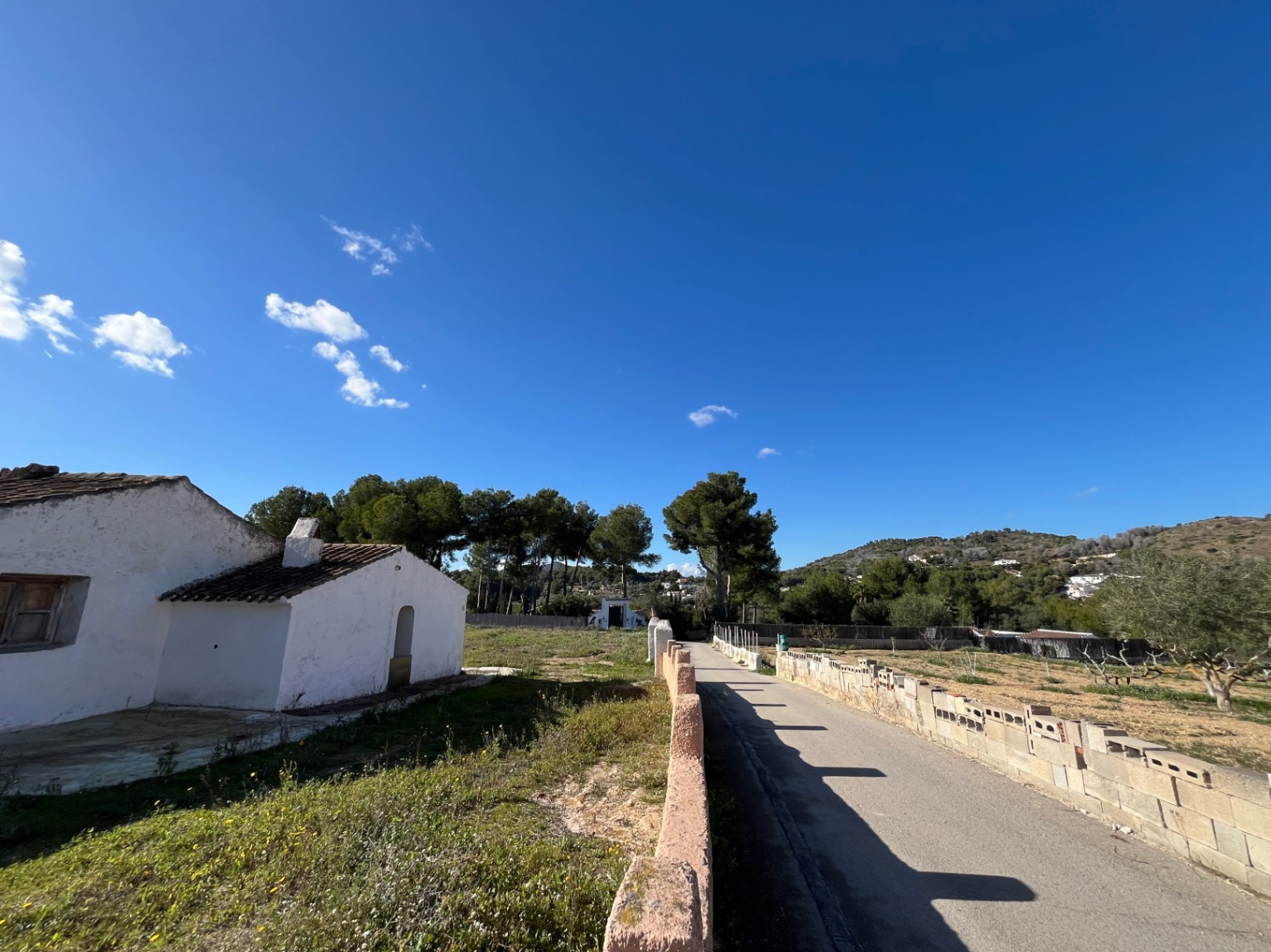 Vista exterior de la finca tradicional con muros blancos, tejado de teja cerámica y camino pavimentado rodeado de pinos y vegetación mediterránea.