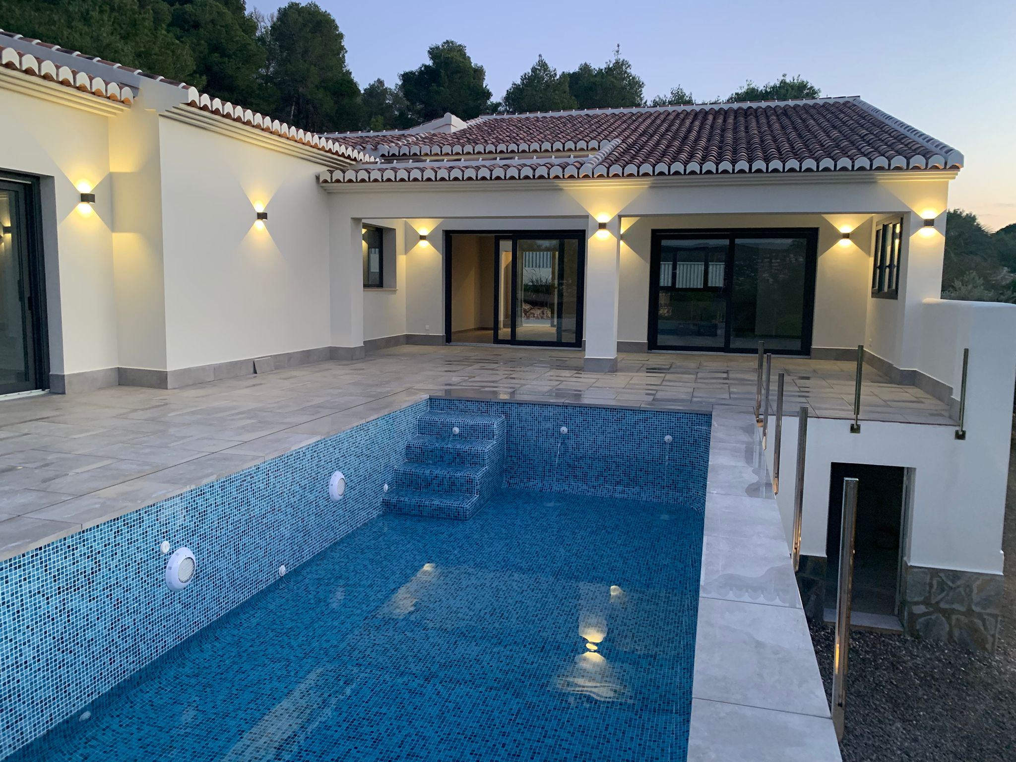 Exterior de villa moderna con piscina de gresite azul, terraza de piedra gris y fachada blanca iluminada con apliques bajo tejado de teja.