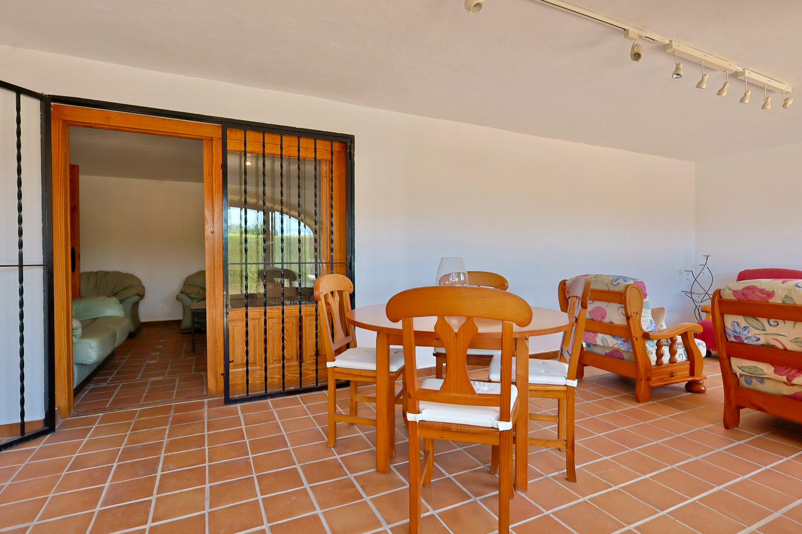 Terraza cubierta con suelo de baldosas de barro cocido, conjunto de comedor de madera y sillones florales. Una puerta corredera con rejas de hierro conecta con el interior.