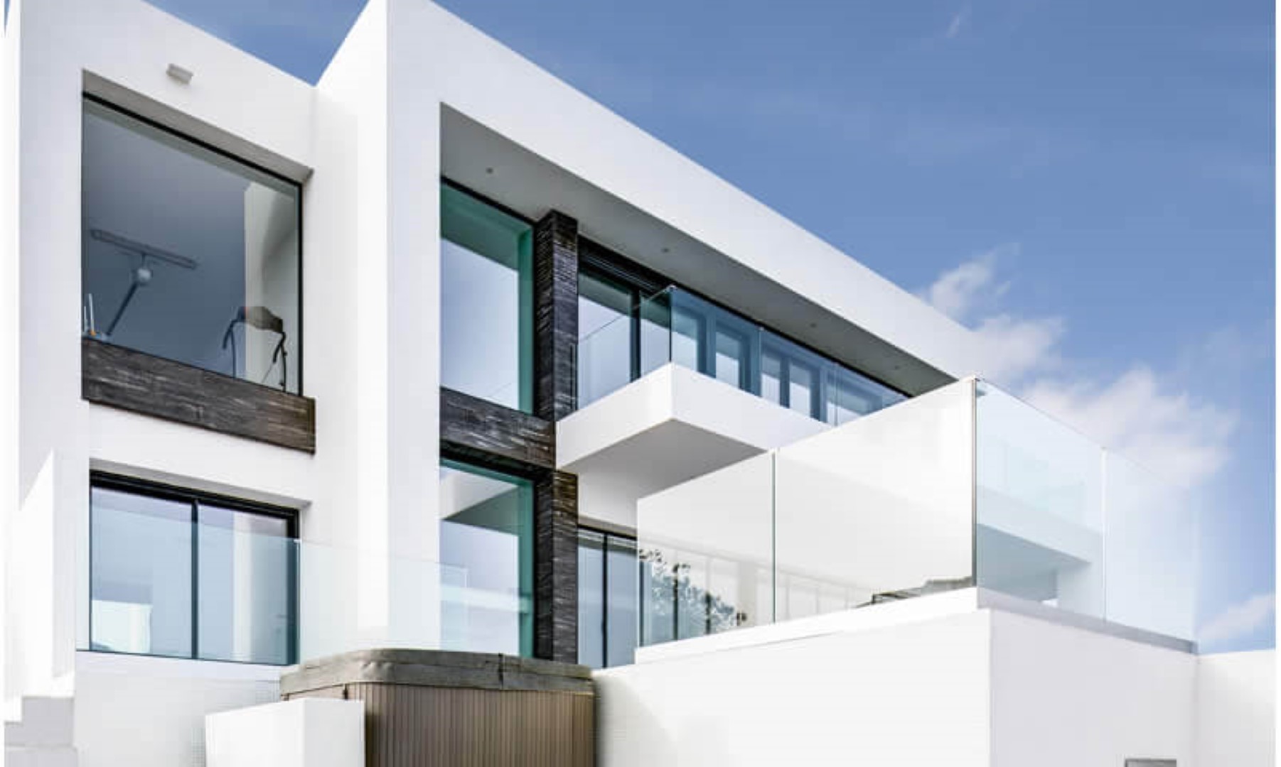 Fachada de diseño geométrico con muros blancos, amplios ventanales y balcones con barandillas de cristal. Destacan los detalles en piedra oscura y una zona de jacuzzi exterior.