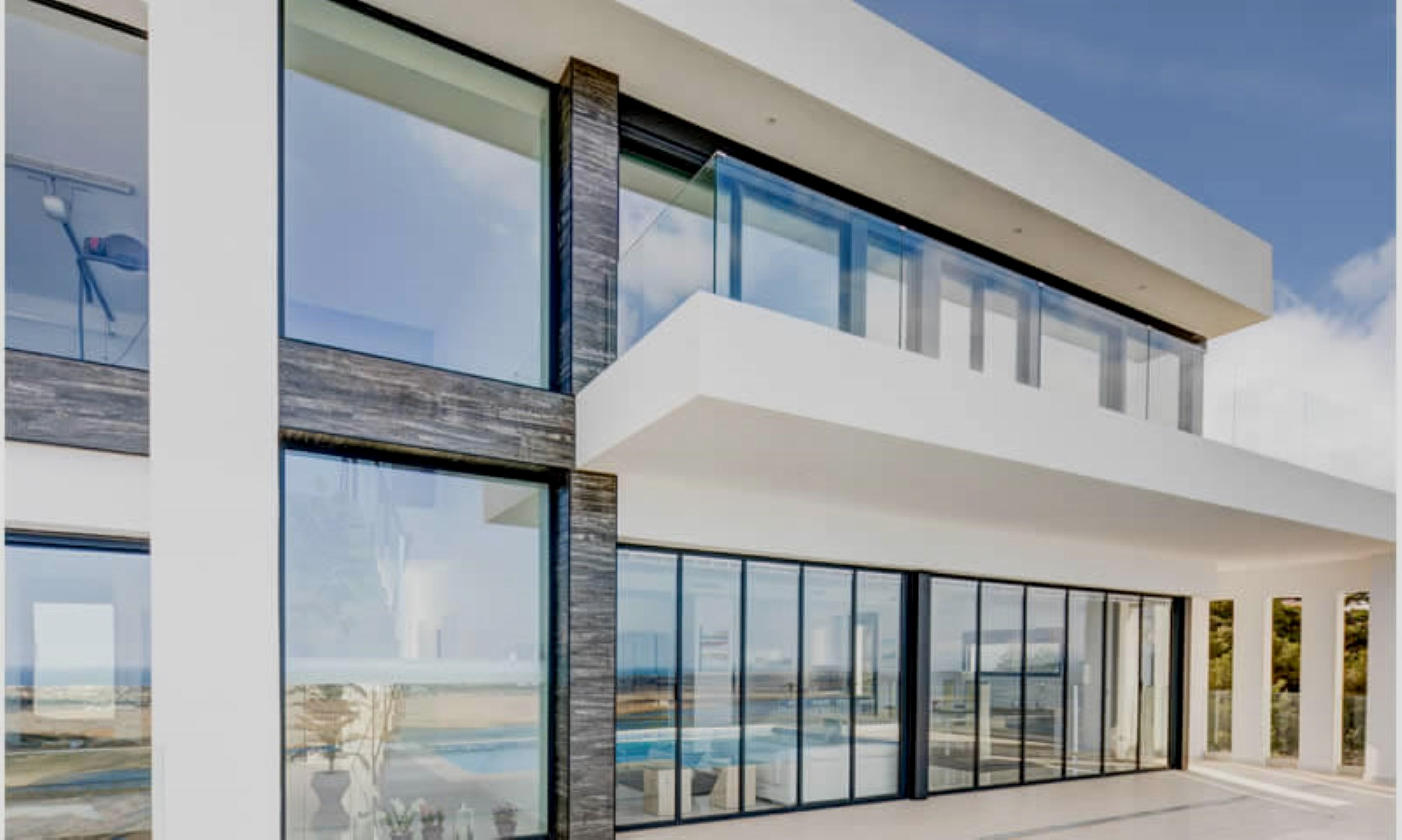 Fachada moderna con grandes ventanales de suelo a techo, detalles en piedra oscura y balcón superior con barandilla de cristal de diseño minimalista.
