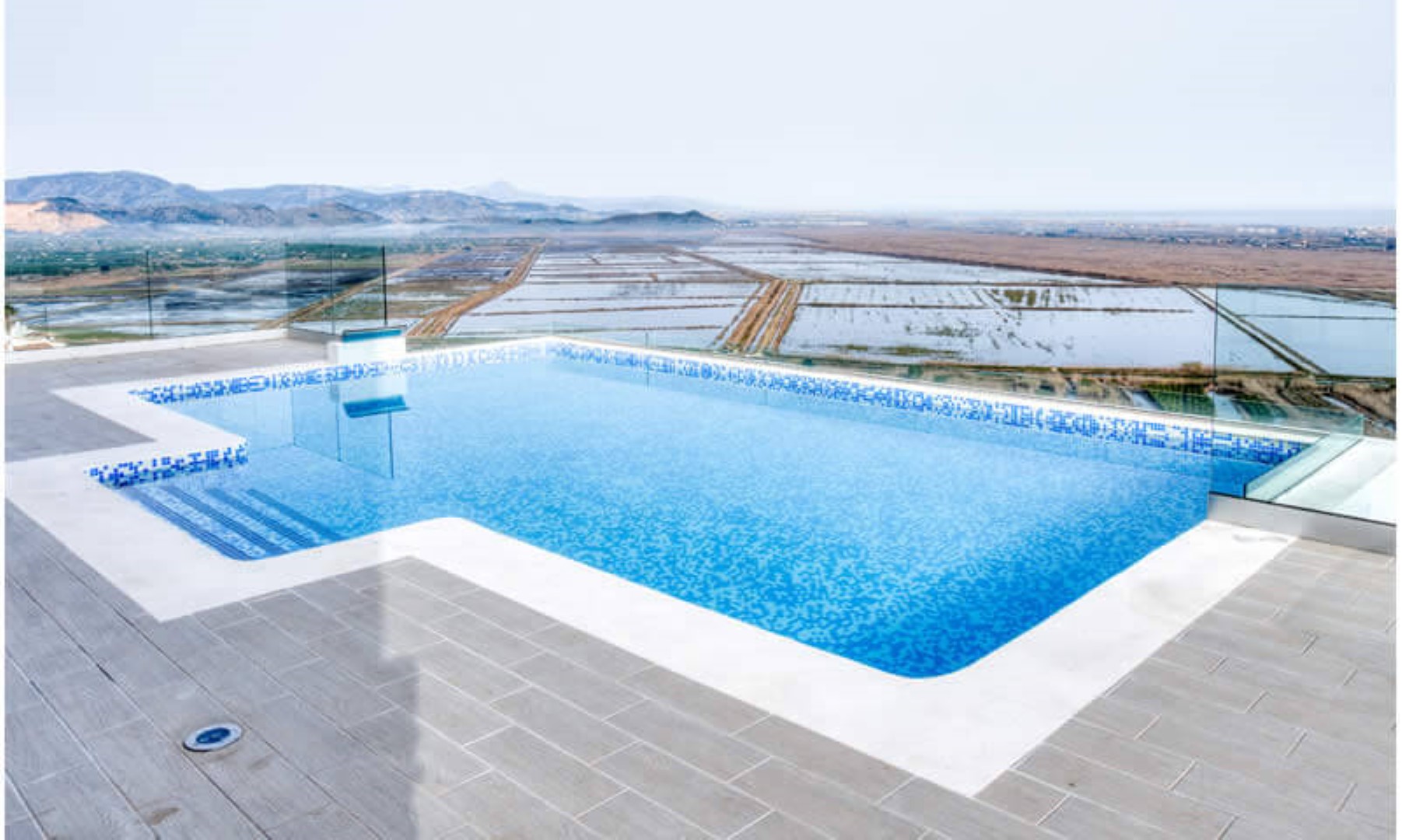 Terraza con piscina infinita de mosaico azul y barandillas de cristal de seguridad, con vistas panorámicas al valle y las montañas circundantes.