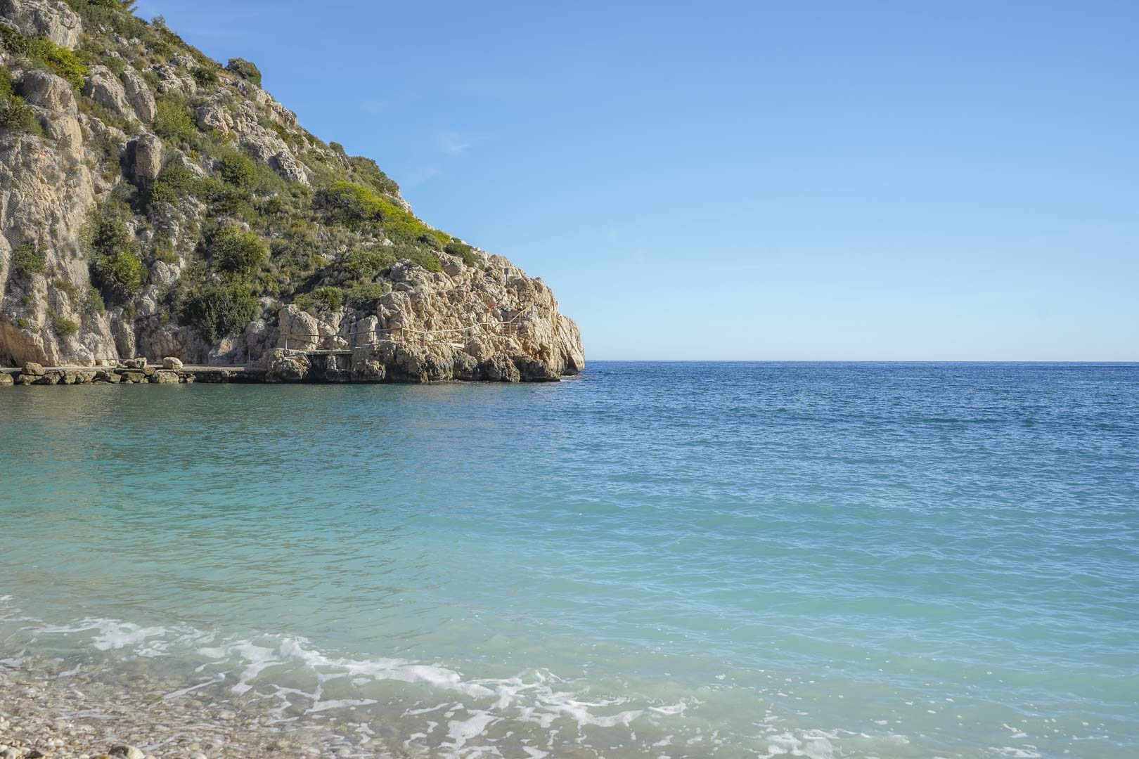 Vista de la Cala de la Granadella en Jávea, destacando sus aguas cristalinas de color turquesa, orilla de cantos rodados y acantilados rocosos con vegetación mediterránea.