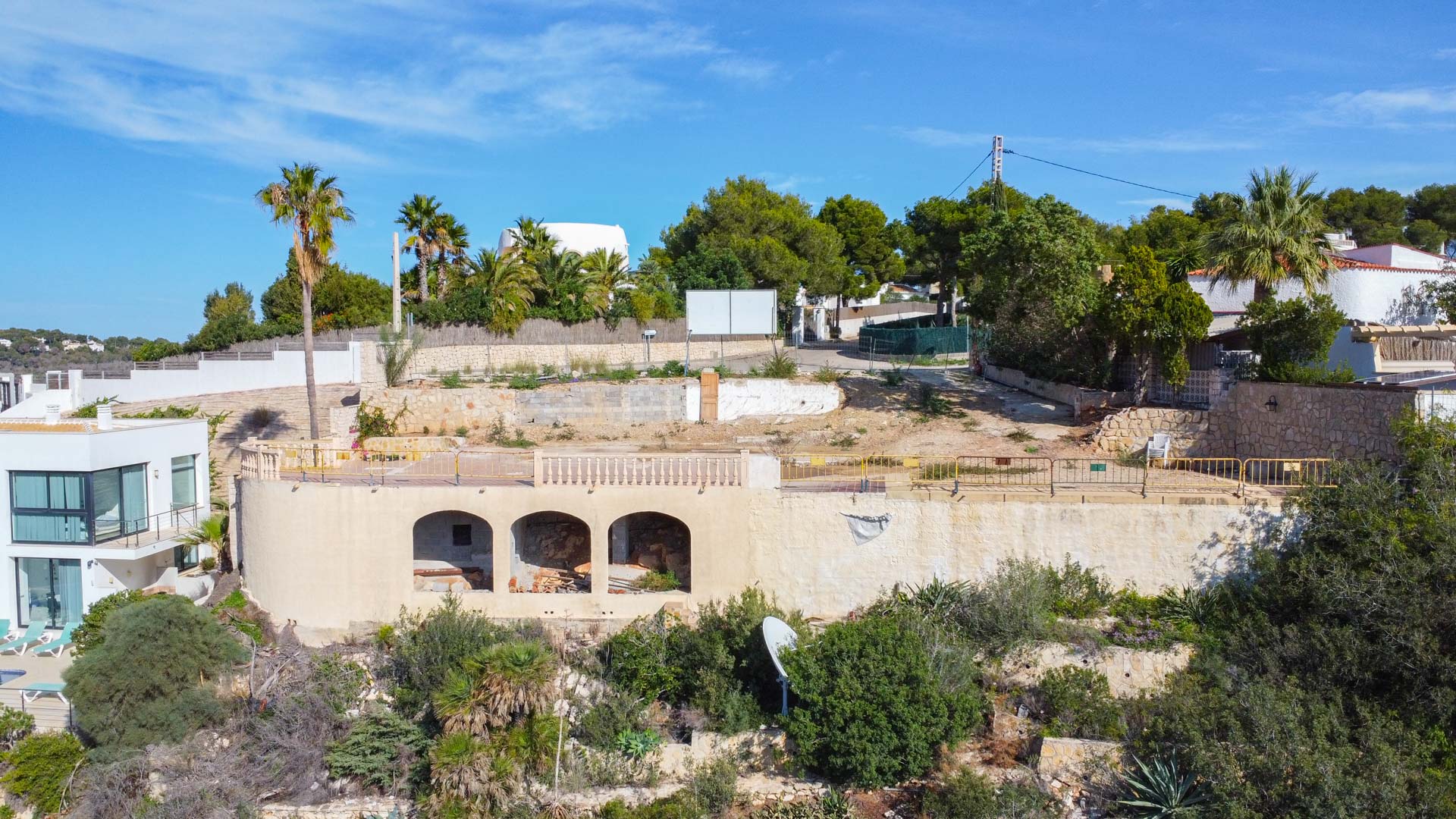 Vista elevada de la parcela en construcción en La Granadella, mostrando el muro de contención con arcos y la plataforma nivelada rodeada de vegetación mediterránea.