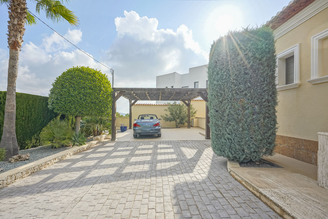 Amplia entrada pavimentada con una pérgola de madera para aparcamiento, rodeada de vegetación mediterránea y muros de piedra natural bajo el sol.