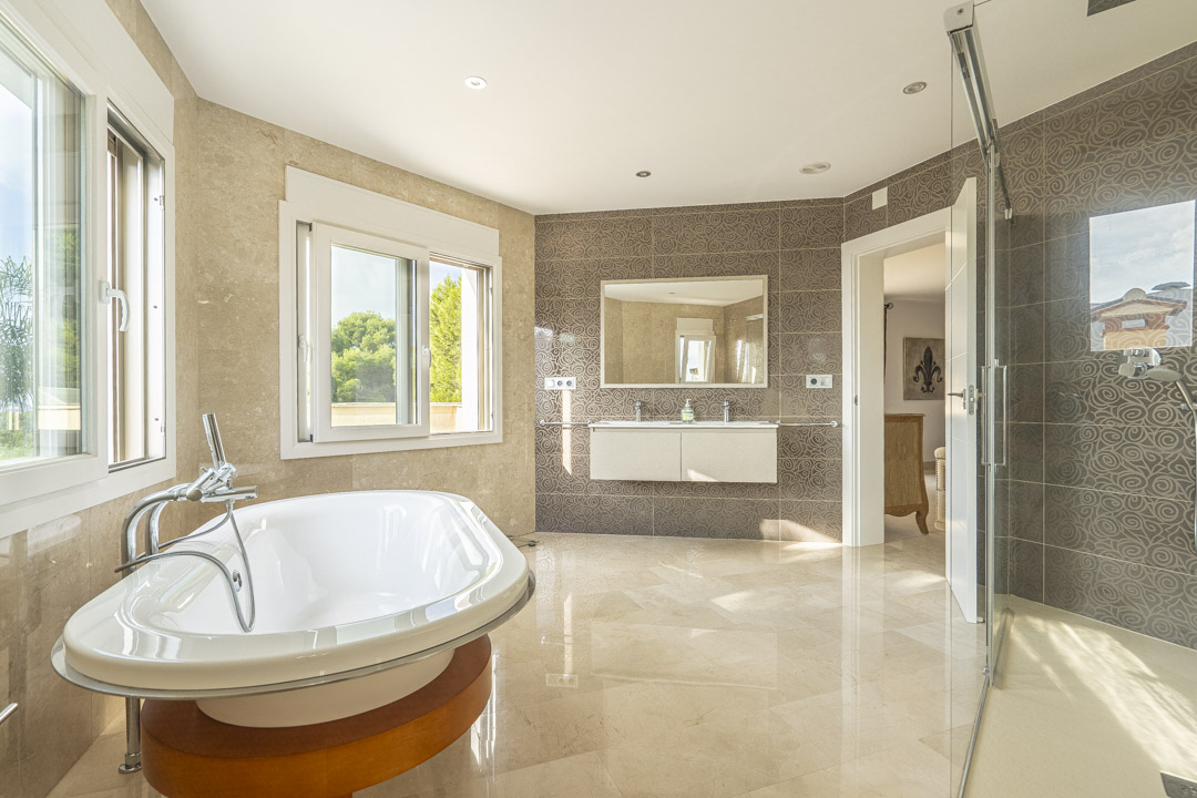 Amplio baño principal con bañera exenta sobre base de madera, lavabo doble suspendido y ducha con mampara de cristal. Destacan los revestimientos de mármol y azulejos decorativos.