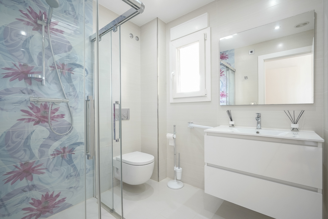 Baño moderno con ducha de obra acristalada, azulejos decorativos de motivos florales, lavabo suspendido blanco y sanitario de pared en tonos neutros.