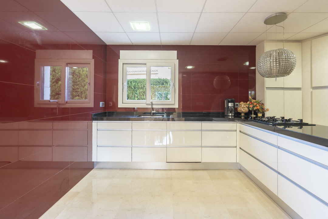 Cocina moderna con armarios blancos sin tiradores, encimera de granito negro y una llamativa pared frontal en rojo brillante. Incluye iluminación empotrada y una lámpara colgante decorativa.