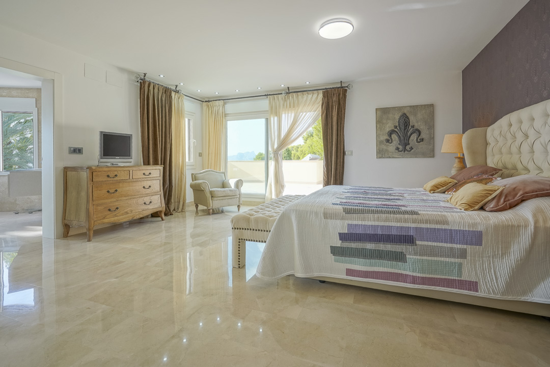 Dormitorio principal con suelos de mármol pulido, cabecero acolchado y acceso directo a una terraza privada con vistas lejanas al Peñón de Ifach.